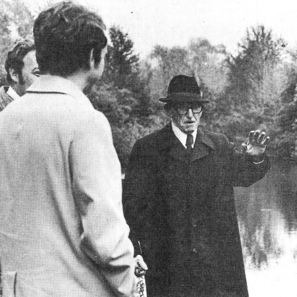 Black and white image from 1976. A man explains to others near a body of water.