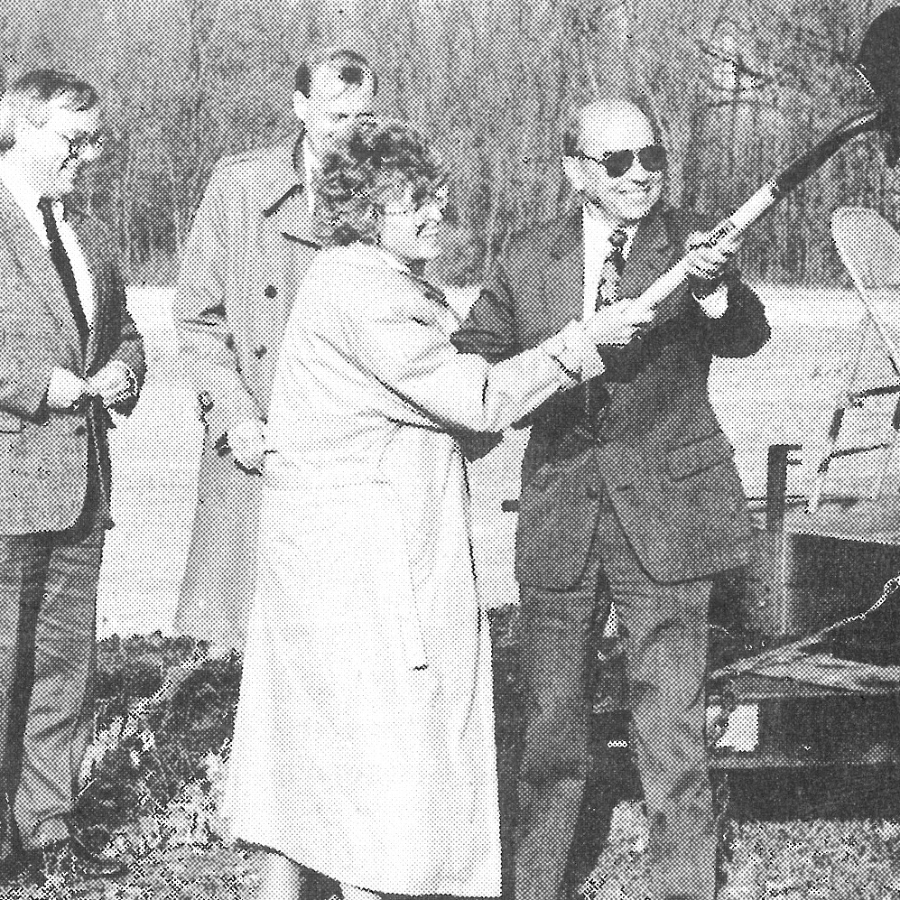 Black-and-white photo from 1991 of people in suits. Woman holds a rifle while a man holds a shovel.