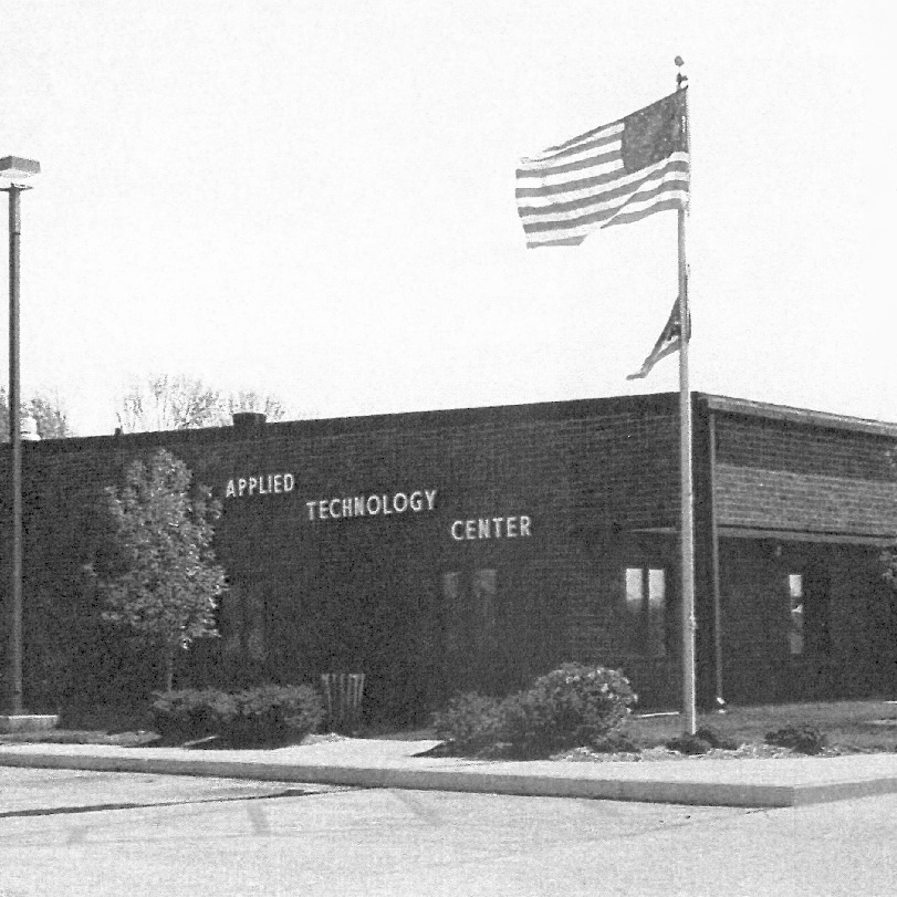 A black-and-white photograph of the Adult Technology building from 1992. A flag flies on a pole in front.