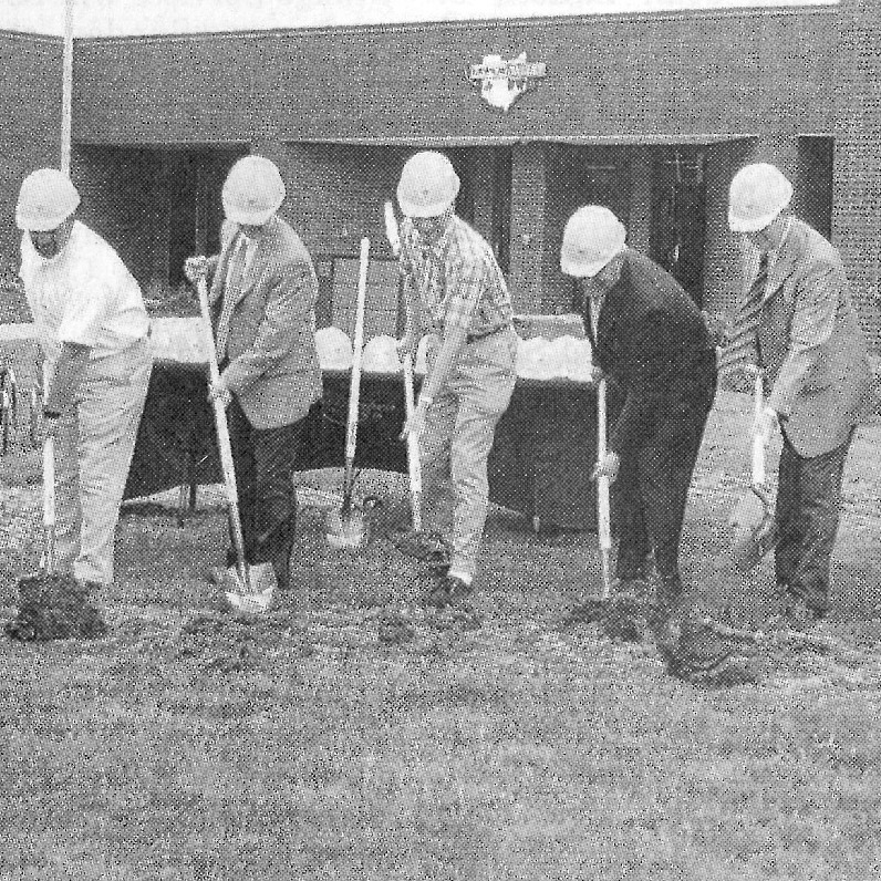 A group of people wearing hard hats and gloves participate in a ground-breaking ceremony for a school building.