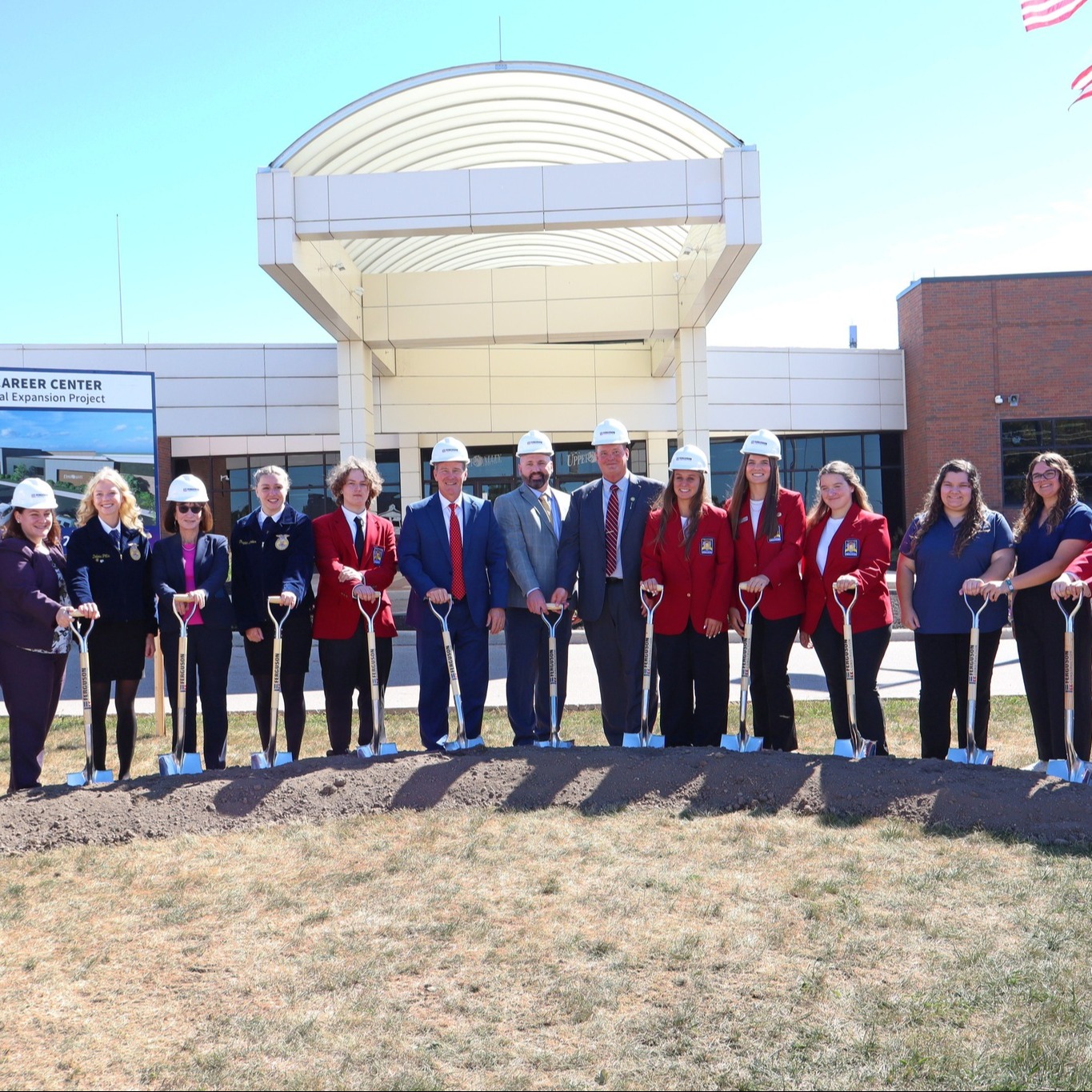 Group of people in hats and formal wear holding shovels in a dirt patch, outside a building.