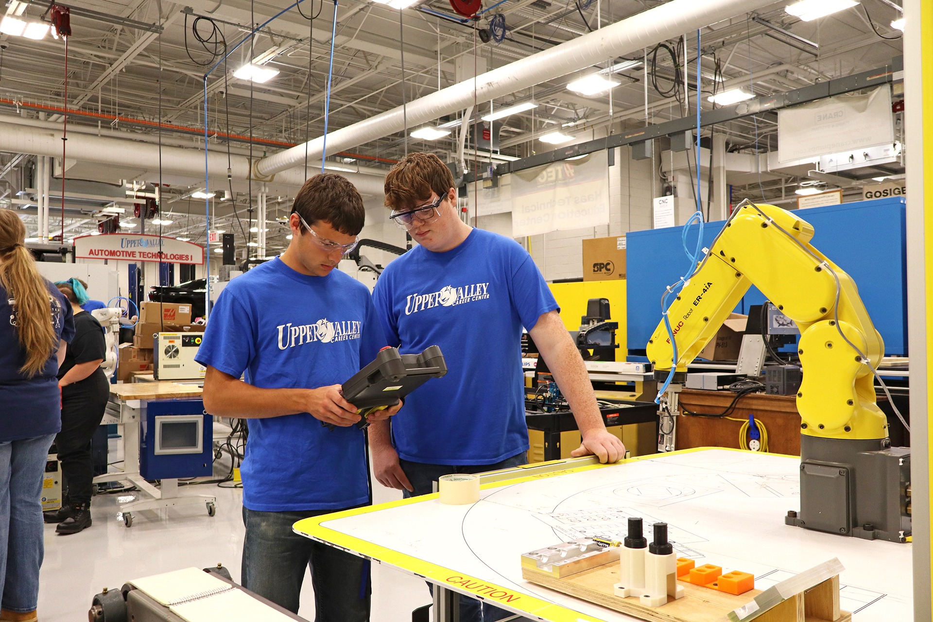 Two individuals in blue shirts and safety glasses in a factory. One holds a device, the other looks over. A yellow robotic arm and white table are nearby.