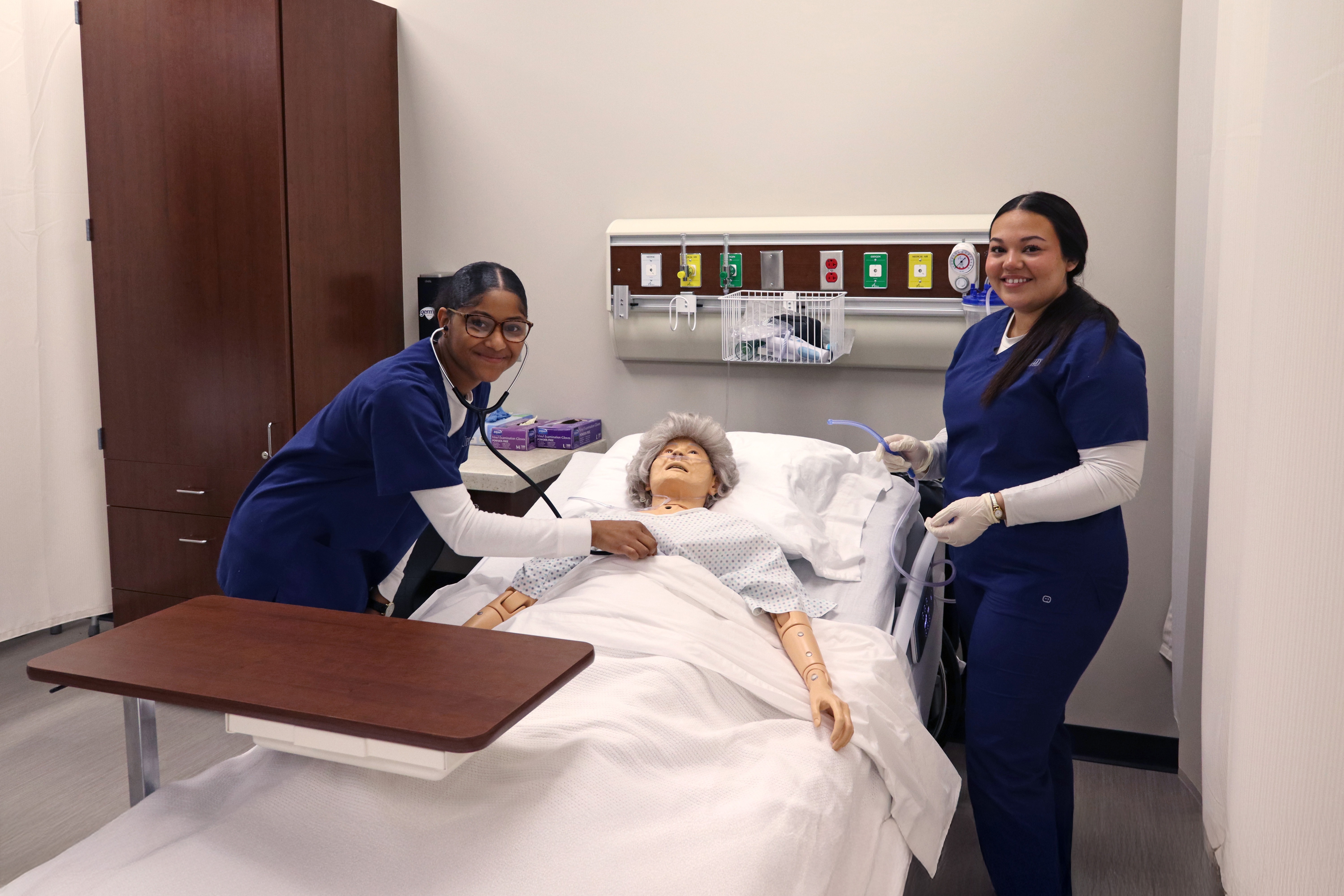 Three women in blue scrubs examine a mannequin on a hospital bed, one holding a stethoscope.