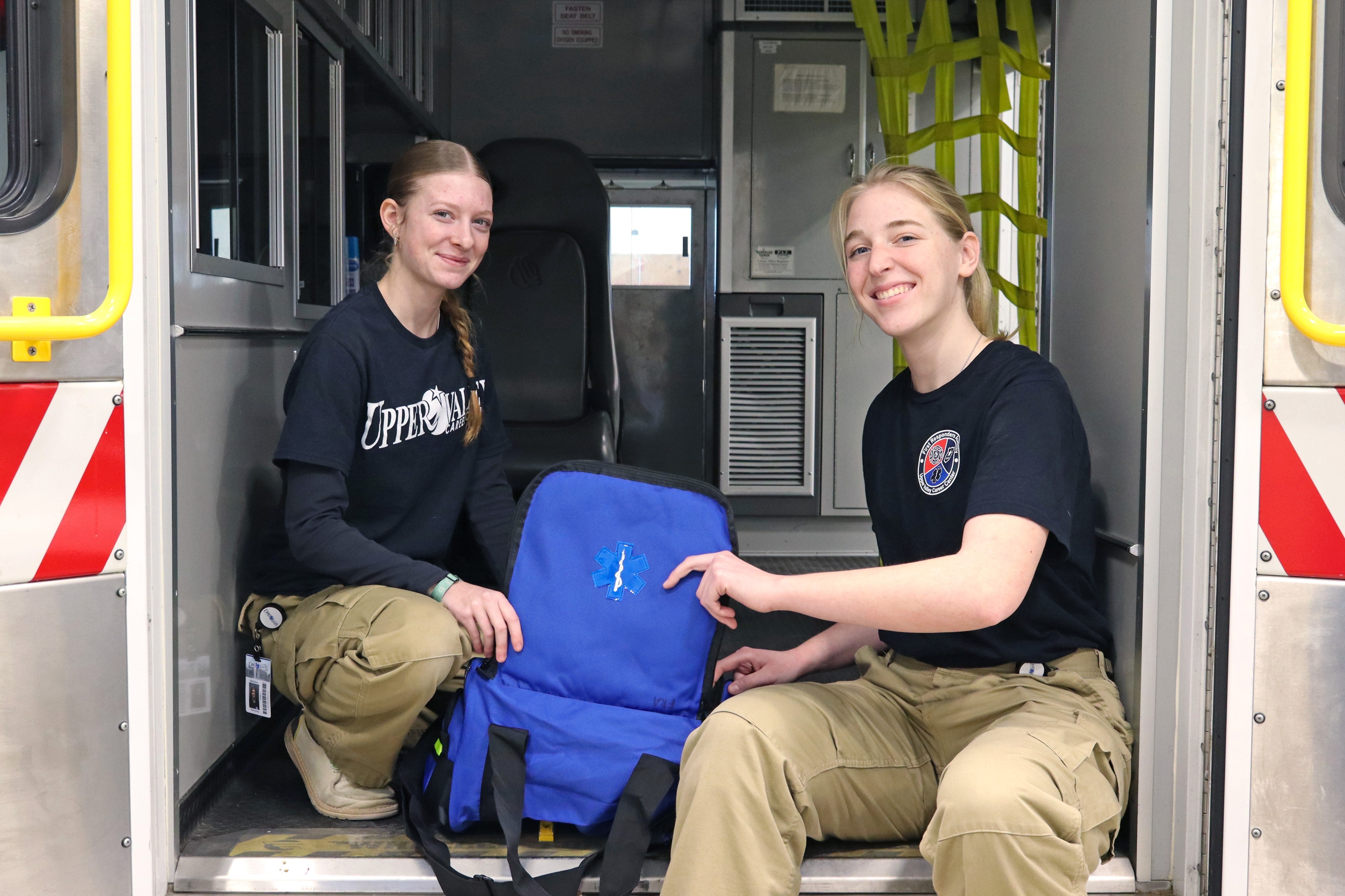 Two women in black t-shirts and khaki pants sit in an ambulance. One holds a blue bag.