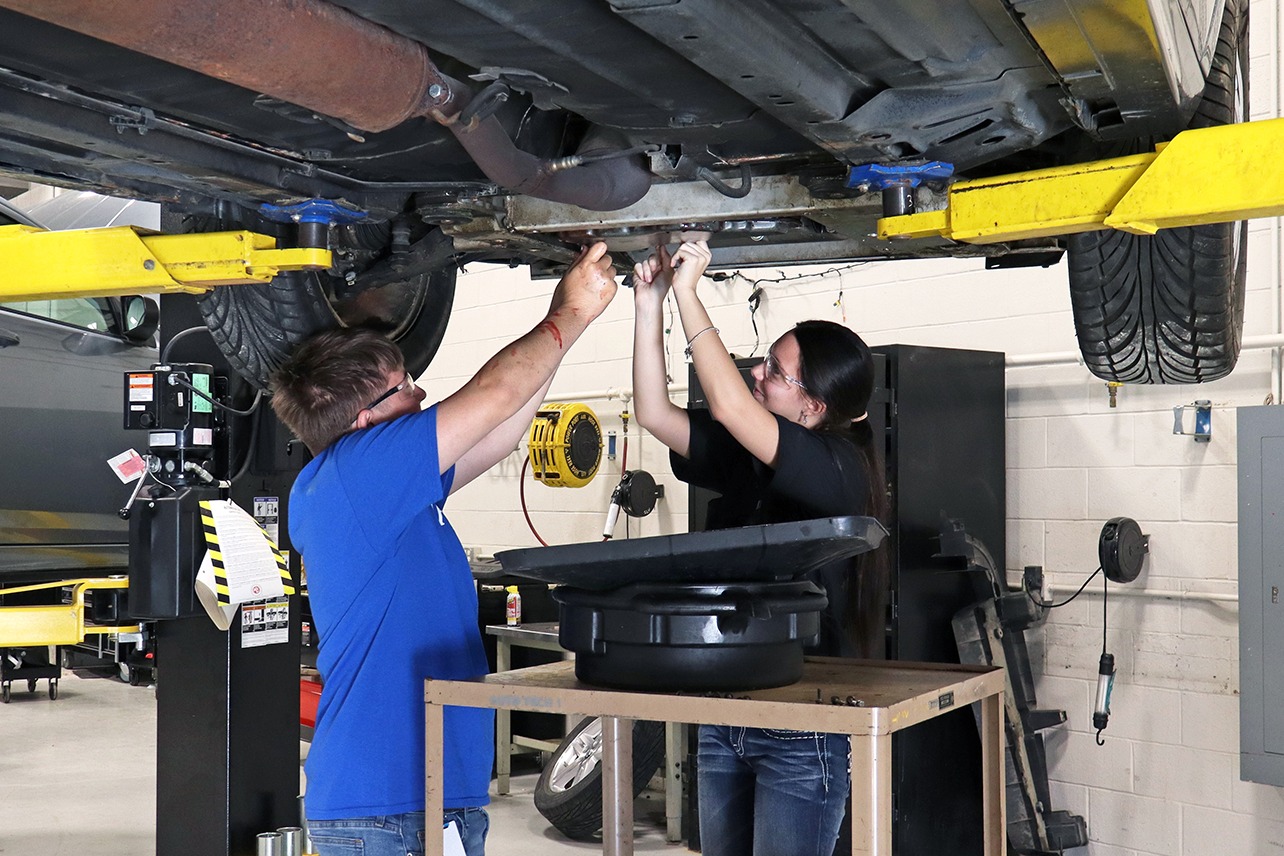 Two people work on a vehicle's undercarriage in a garage with yellow lift arms and gray electrical panels.