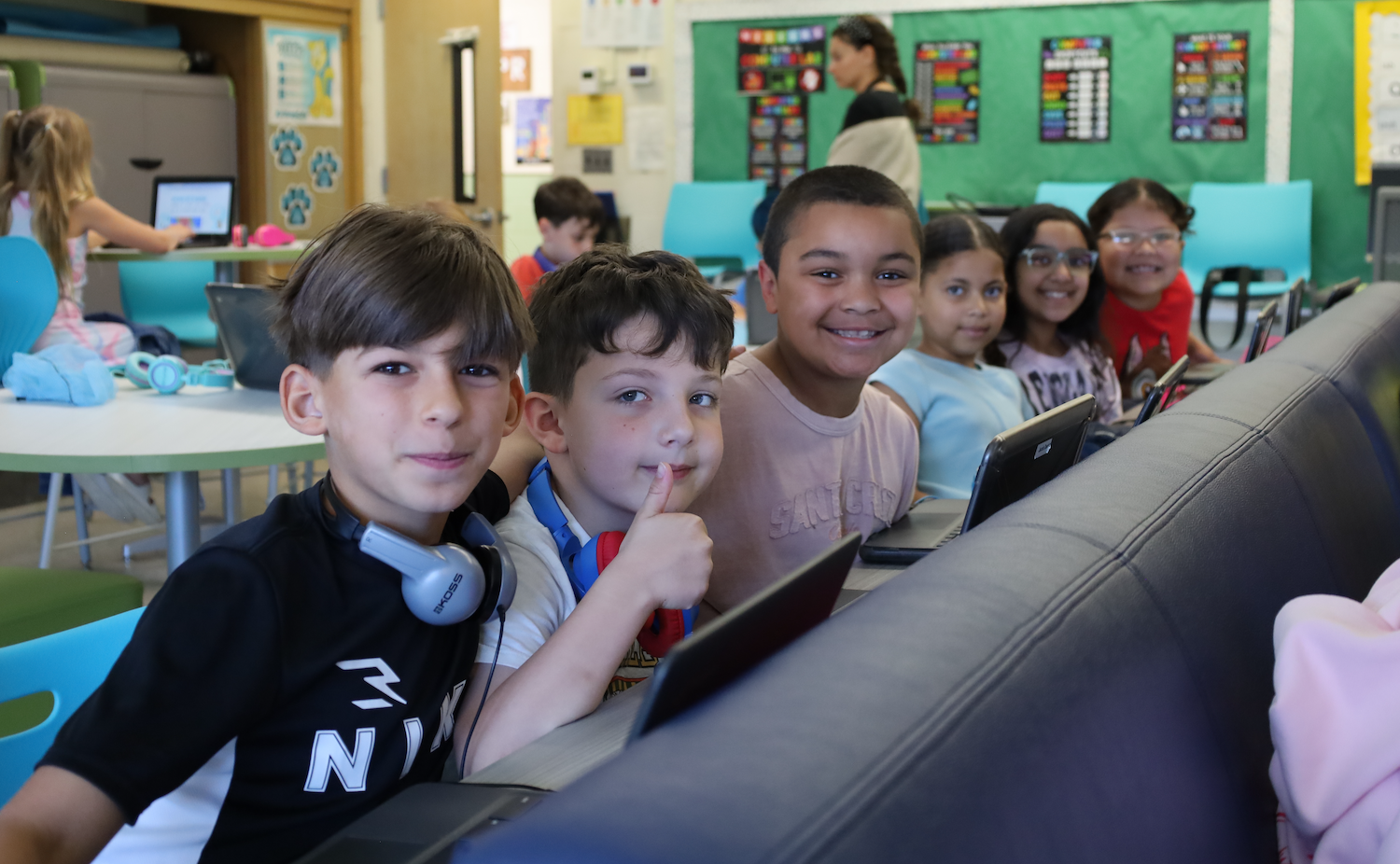 Several children seated around computers in a classroom. Two boys in front wearing headphones and one boy making a peace sign.
