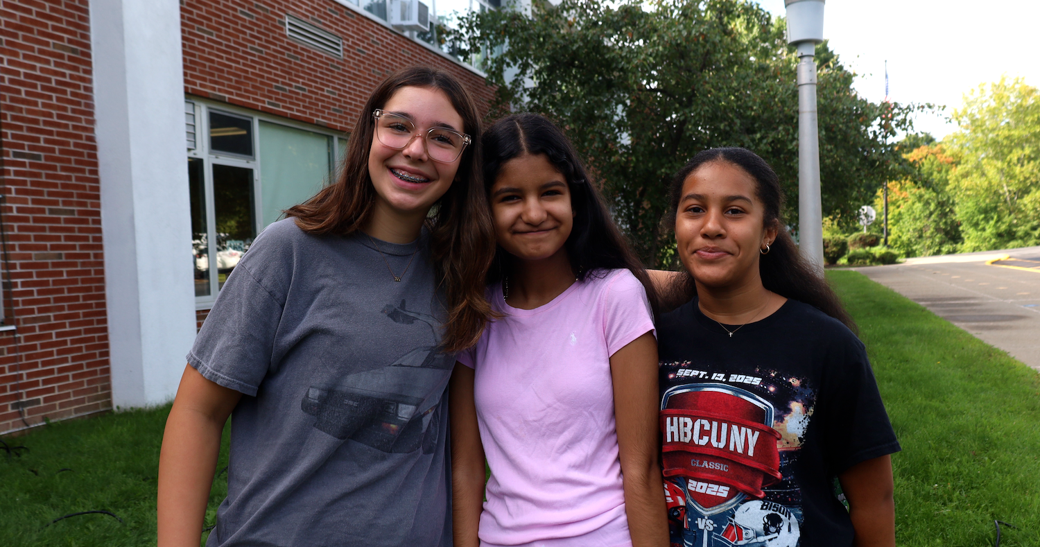 Three girls stand together on a lawn with a brick building and trees in the background.