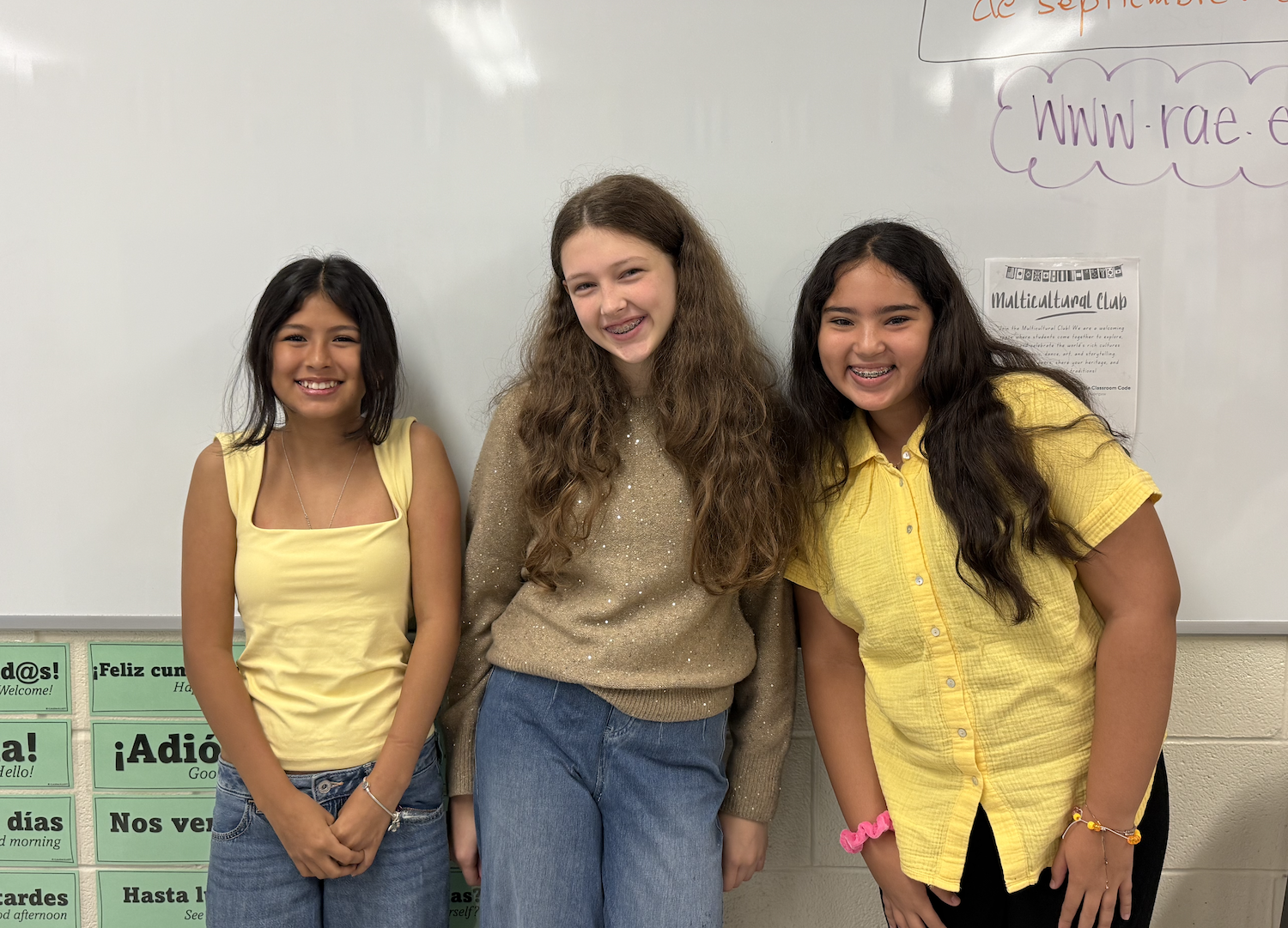 Three young females stand next to each other in front of a whiteboard. They wear yellow tops.