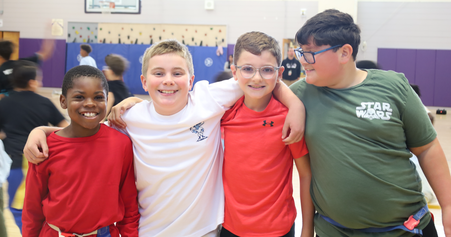 Four kids in a gym, two in red shirts and two in white and green shirts, smiling and posing for a photo.