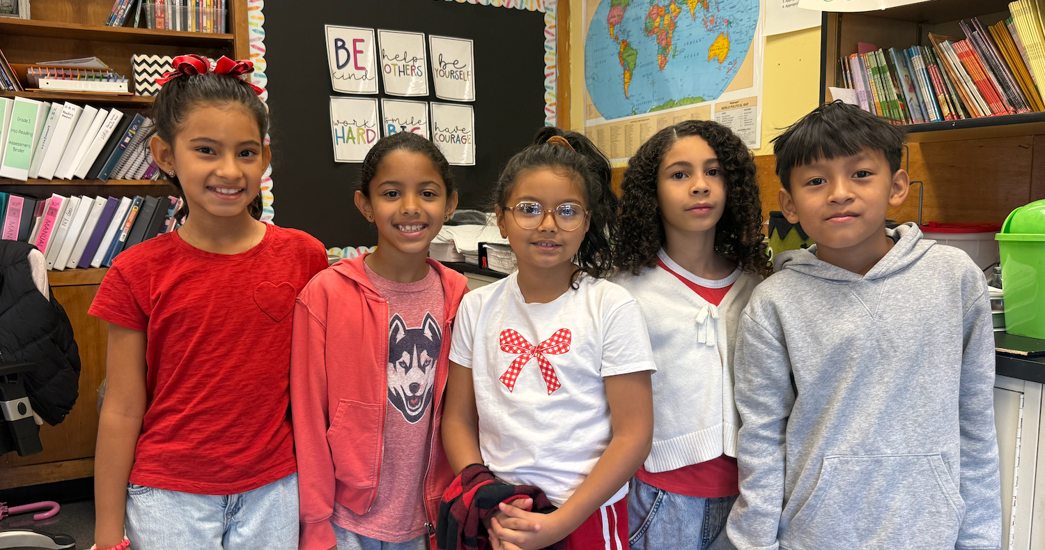 Five children, two girls and three boys, stand in a classroom. One girl wears a red shirt, another a pink jacket. A girl has a white top.