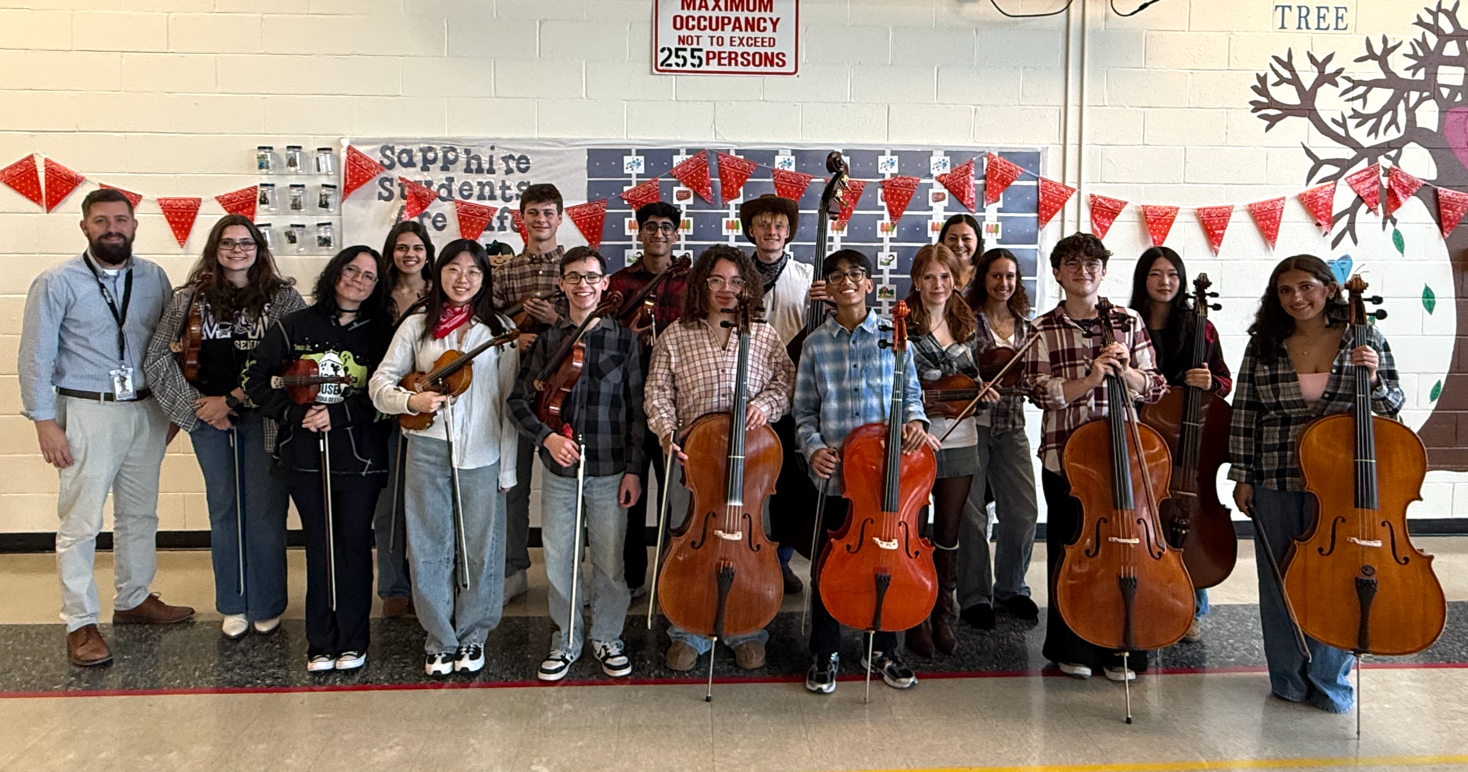 People standing with cellos and violins in a room with a decorated wall.