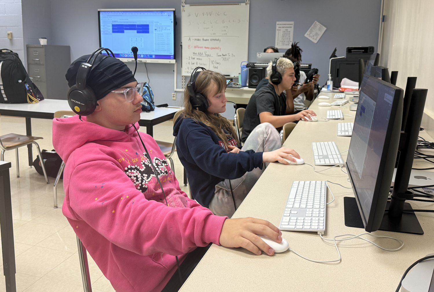 Several students wearing headphones, seated at computers with keyboards and mouse, with a whiteboard in the background.