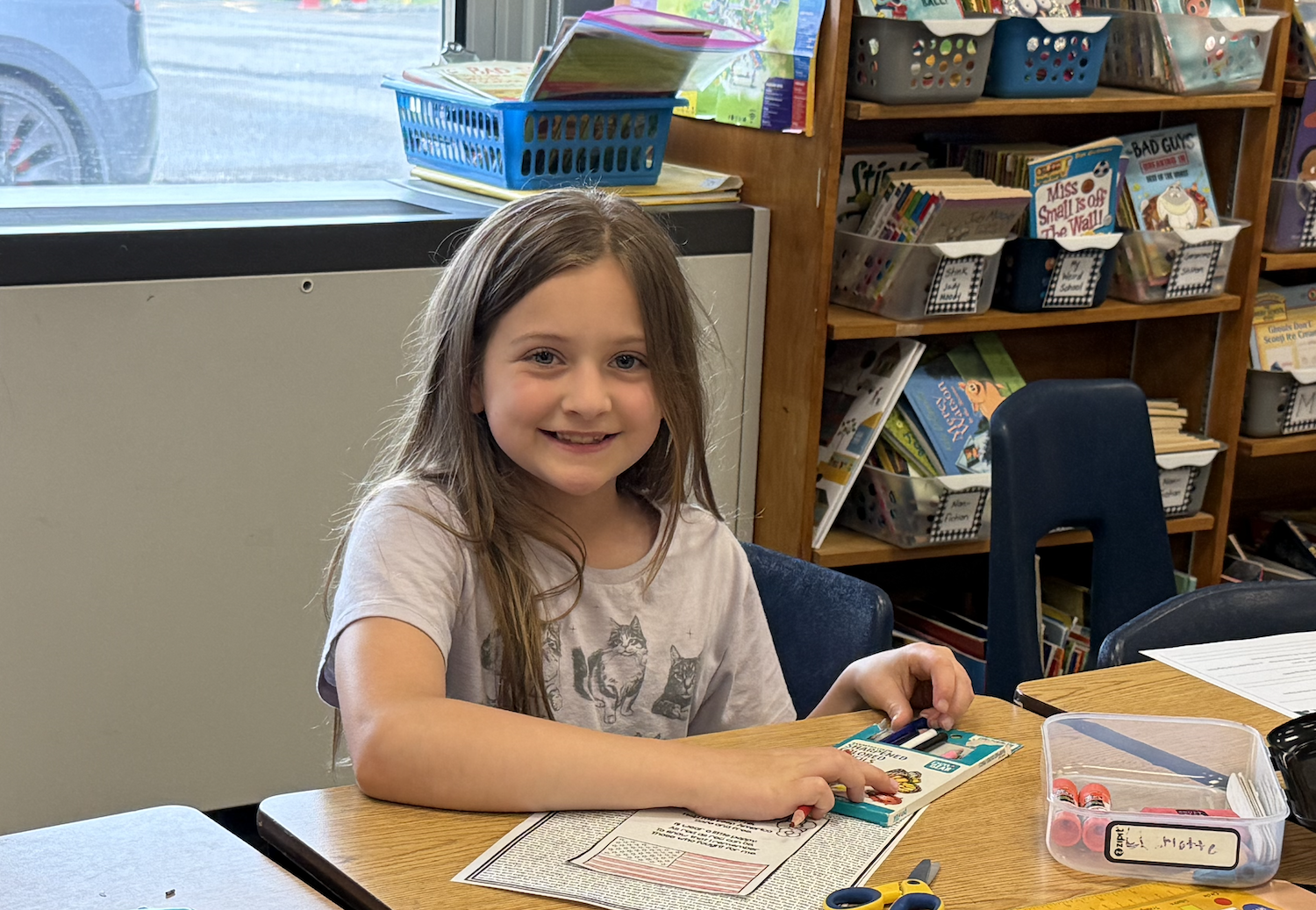 A girl sits at a desk in a classroom, smiling, and working on a puzzle. Behind her is a wooden bookshelf filled with books and supplies.