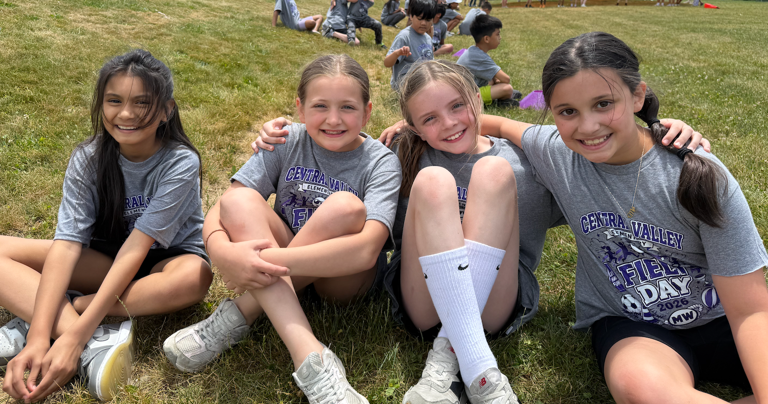 Four girls in gray t-shirts sit on grass, smiling at the camera. One wears white socks, and others have ponytails.
