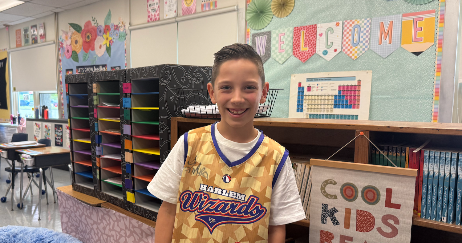A boy with short hair smiles in a classroom. He wears a gold jersey and stands by a bookshelf.