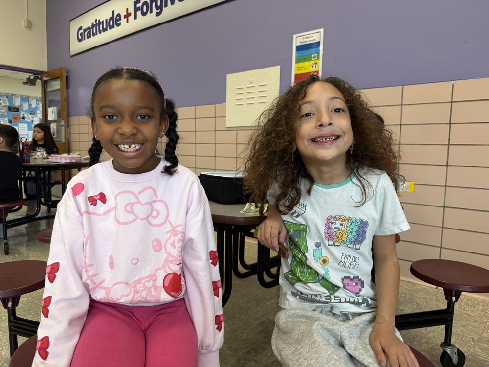 Two girls sit on stools in a classroom, smiling at the camera. Behind them is a wall with a bulletin board.