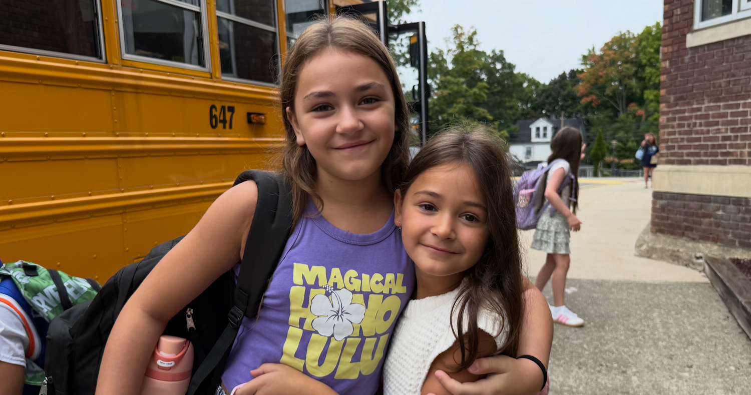 Two girls embrace, one wearing a purple "MAGICAL LUPA" shirt. A school bus is in the background.