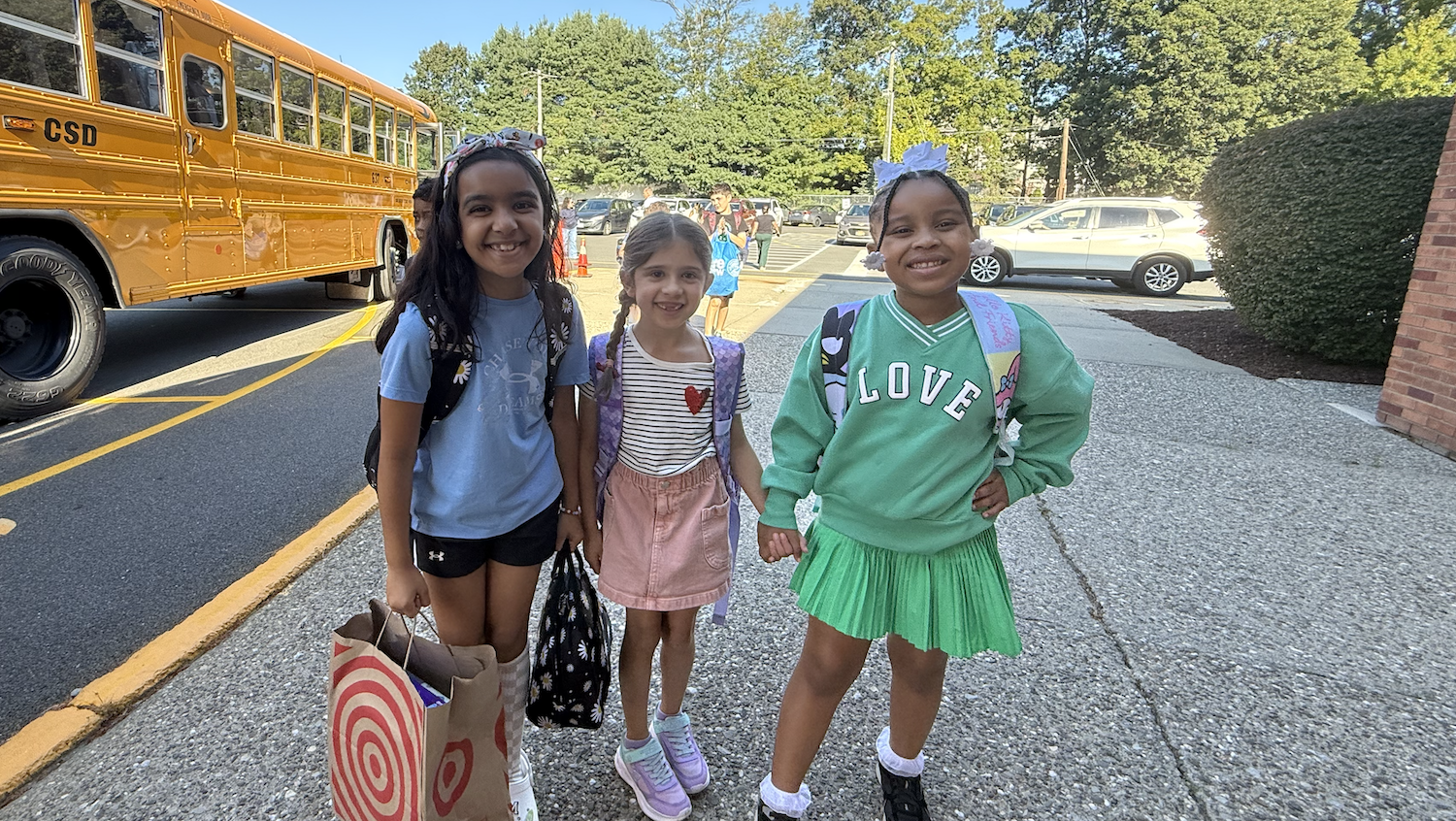 three girls, wearing green, pink and blue in front of a school bus