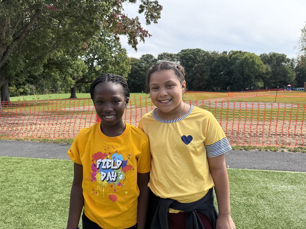 Two girls in yellow shirts stand in a park. Behind them is a fence and trees.