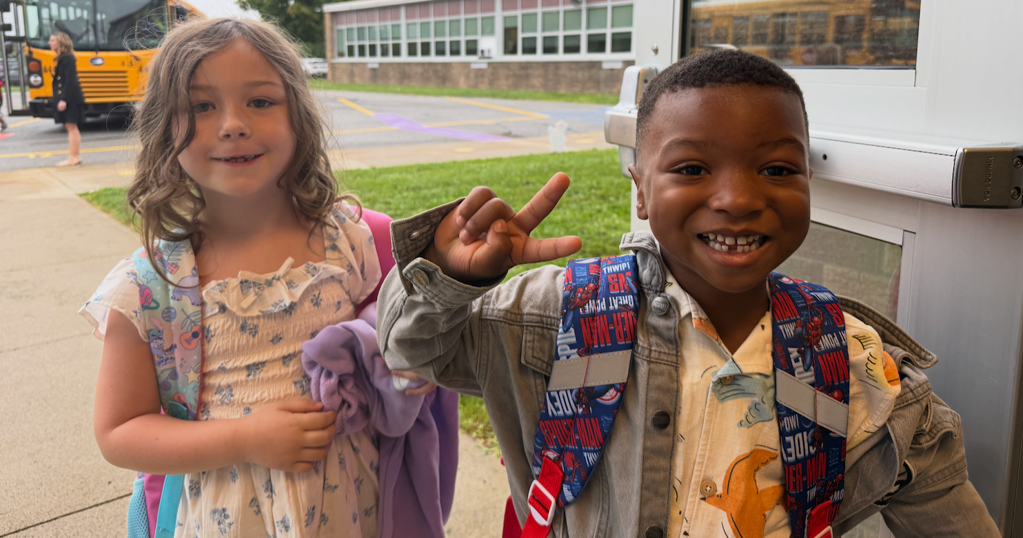 Two kids smiling at the camera, one with a backpack, the other making a peace sign, on a sidewalk.