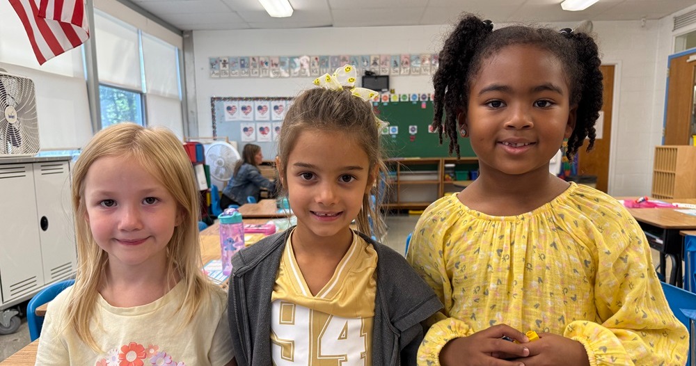 Three young girls in a classroom with yellow shirts. The middle girl wears a jacket. The background shows a desk and an American flag.