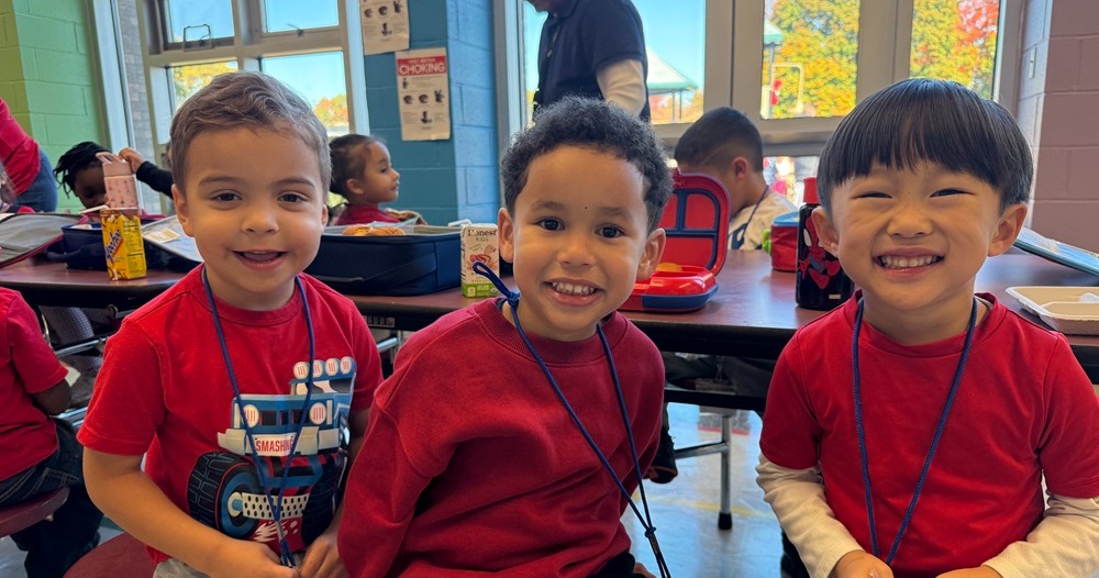 Three children in red shirts sit at a table. A man in black stands near a door.