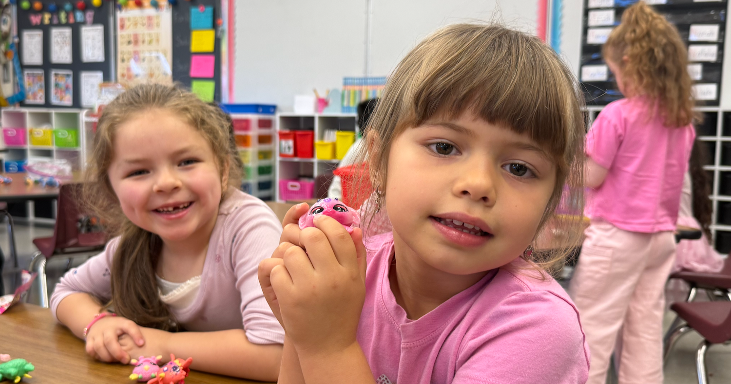 Two girls sitting at a table, one holding a pink toy, smiling and looking at it.