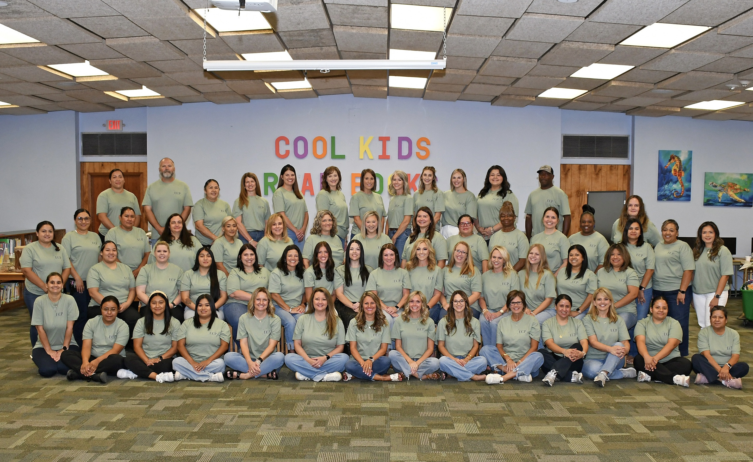 A group of people in green shirts, posing for a photo in a room with a "Cool Kids" mural.