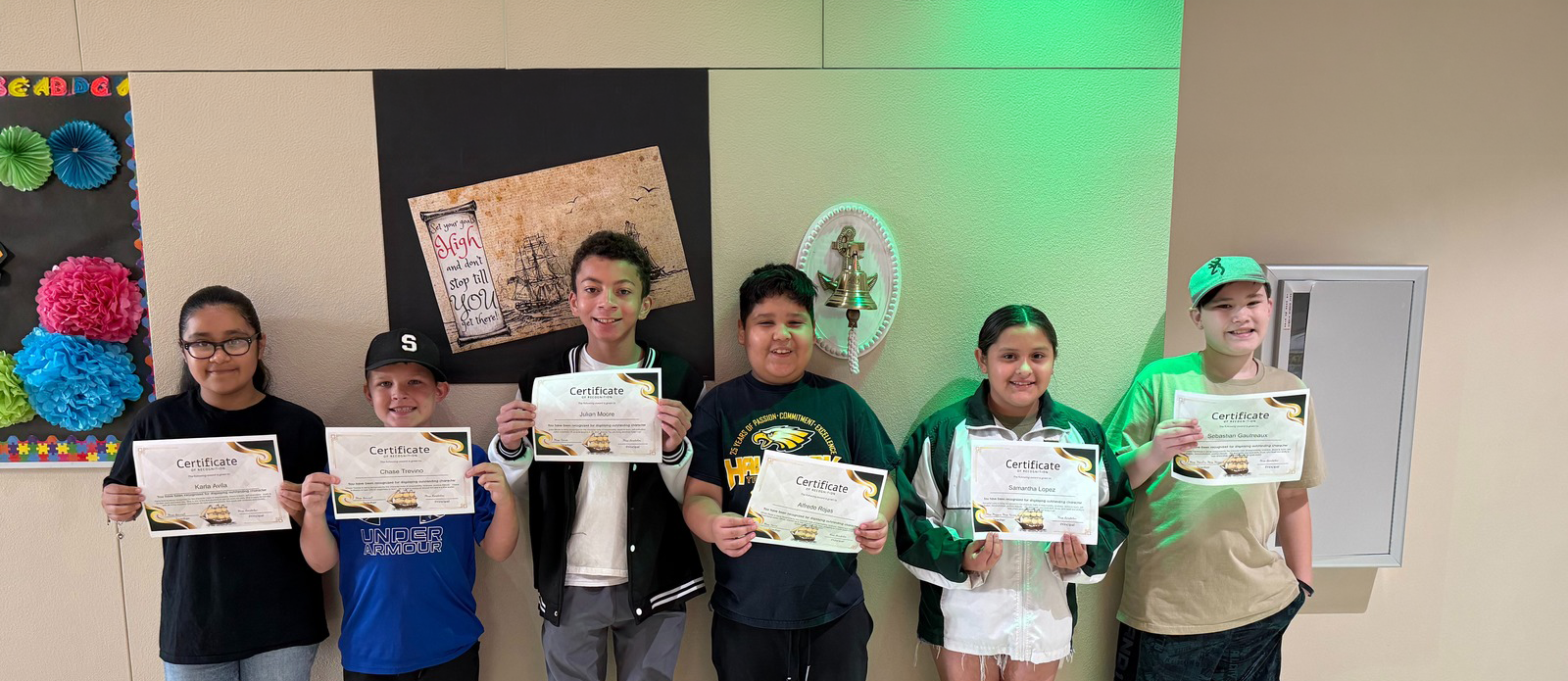 Seven students stand in a row holding certificates. Behind them, a bulletin board displays colorful decorations.