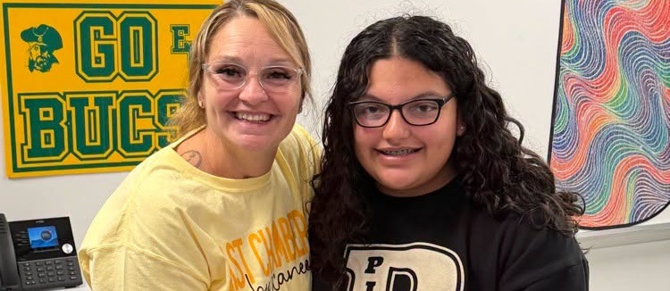 Two women in a classroom. One holds a trophy and the other wears glasses. Behind them is a yellow sign.