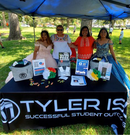 4 women stand behind a table with Tyler ISD tablecloth and flyers/pamphlets on the table. Two of the young ladies are wearing full evening gowns