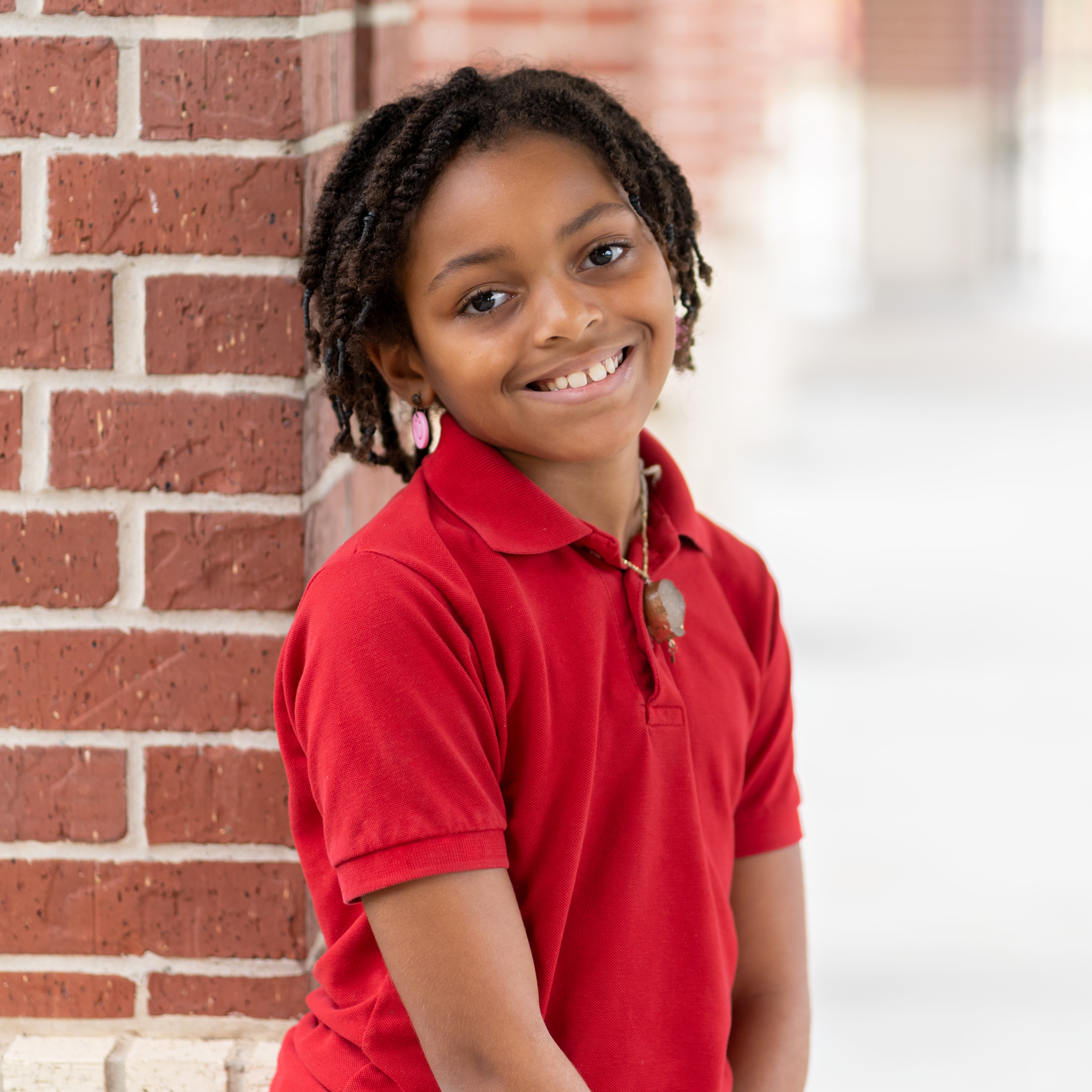elementary age African-American girl wearing a red polo shirt leaning against a brick wall