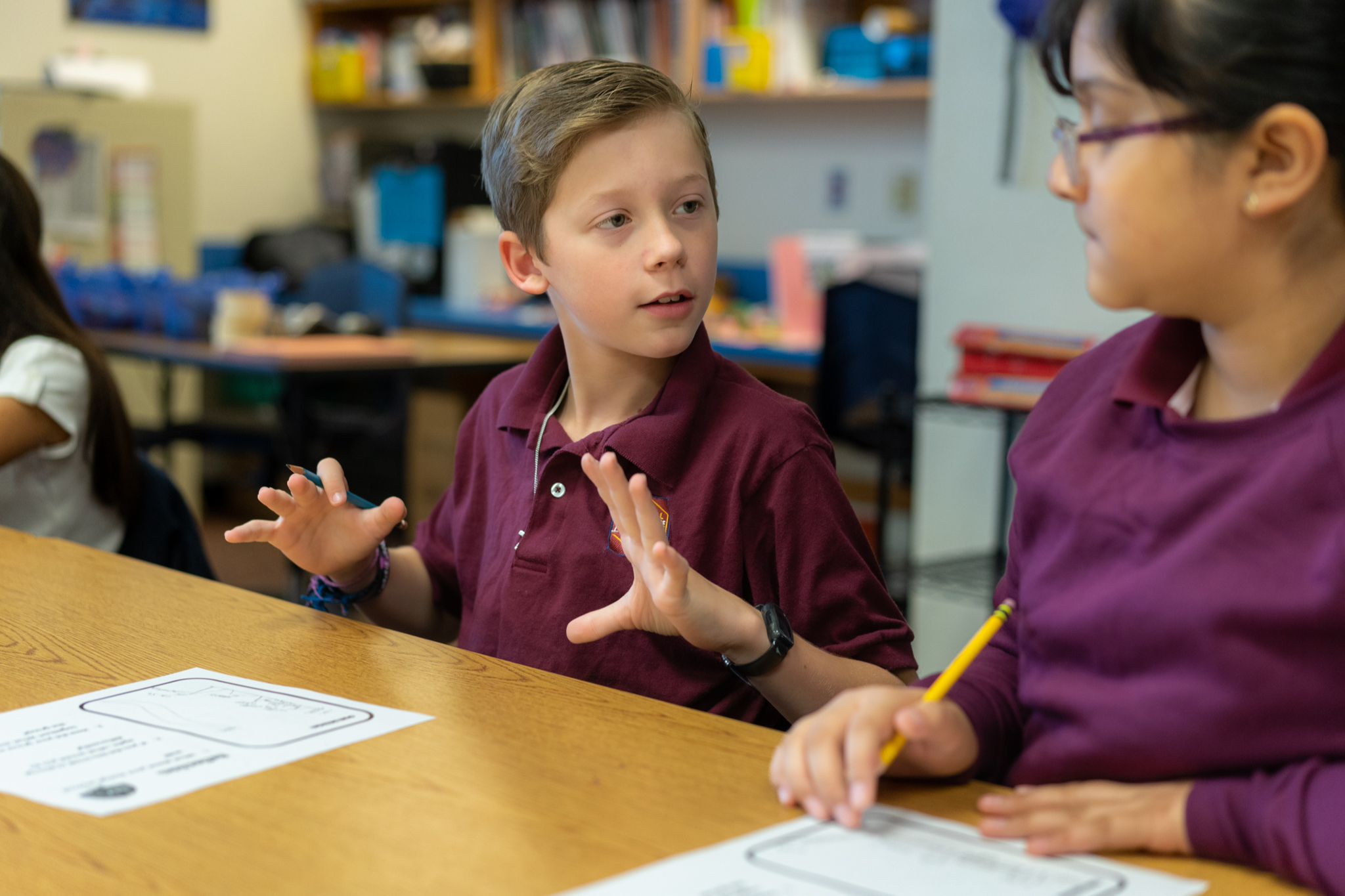 two elementary age students wearing a maroon polo shirt sitting at a desk with papers in front of them