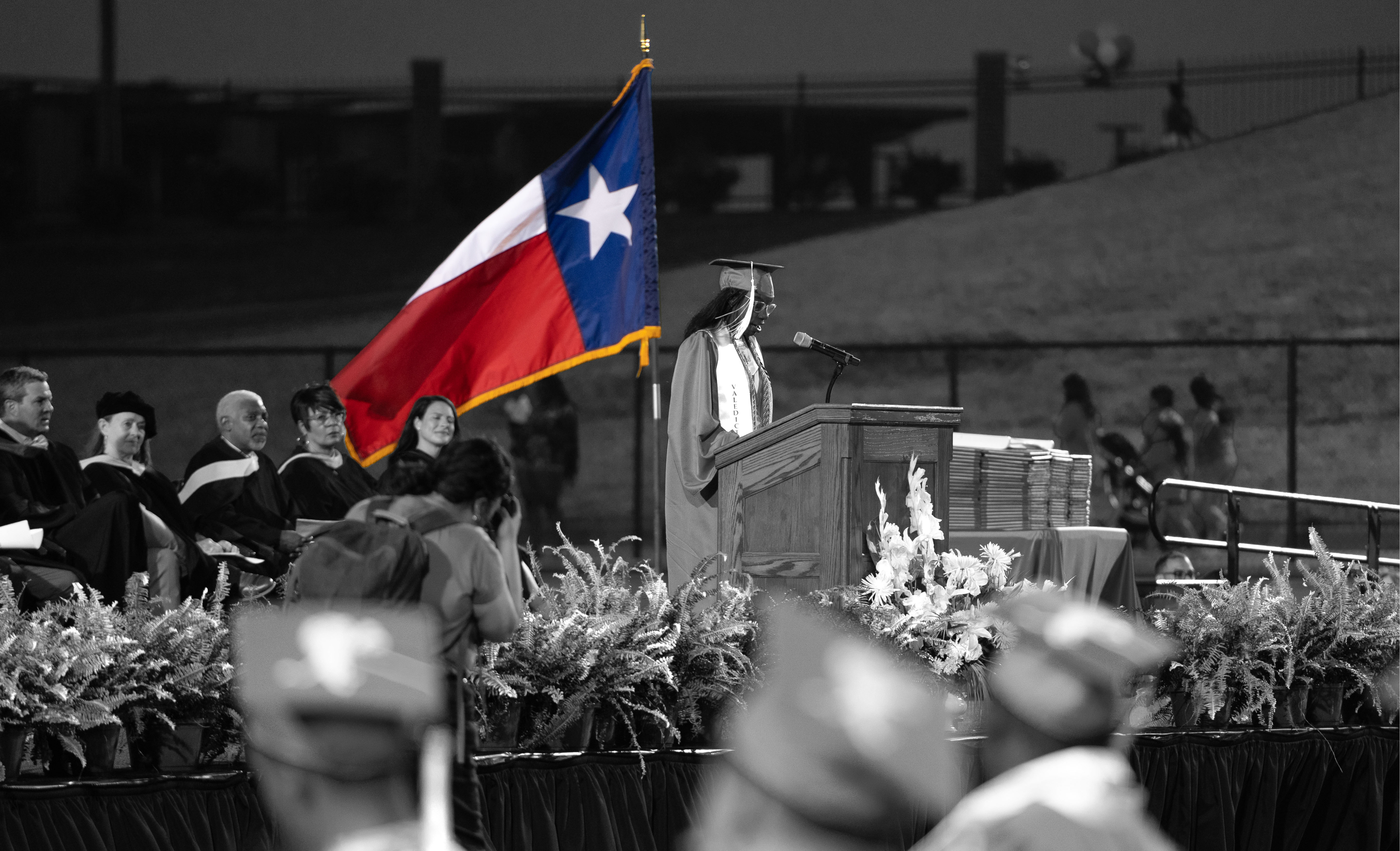 black and white photo of people sitting on stage in their graduation regalia, speaker in cap and gown with Texas flag (in color) flying behind her