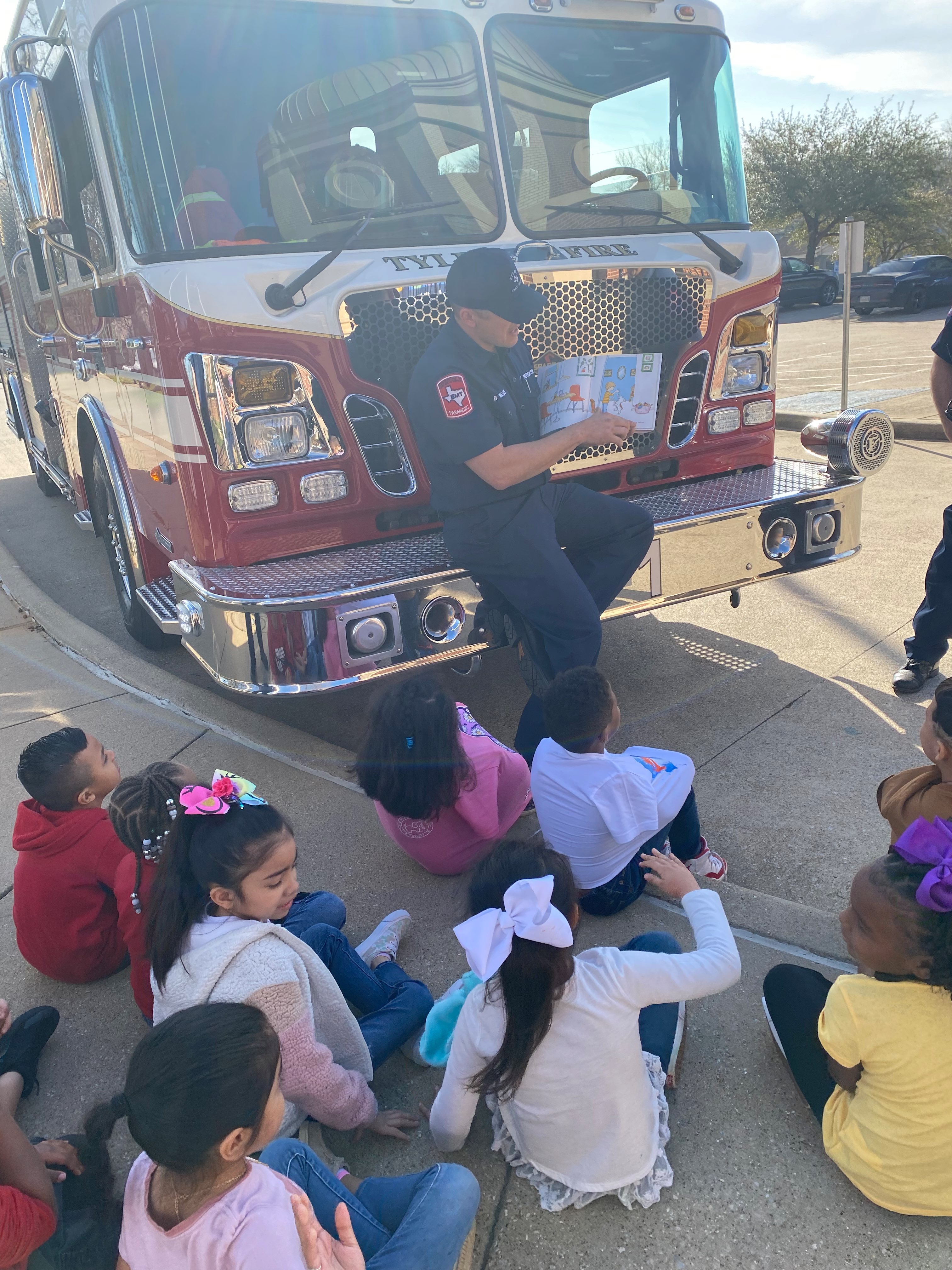 fireman sitting on front of his truck reading a book to kids