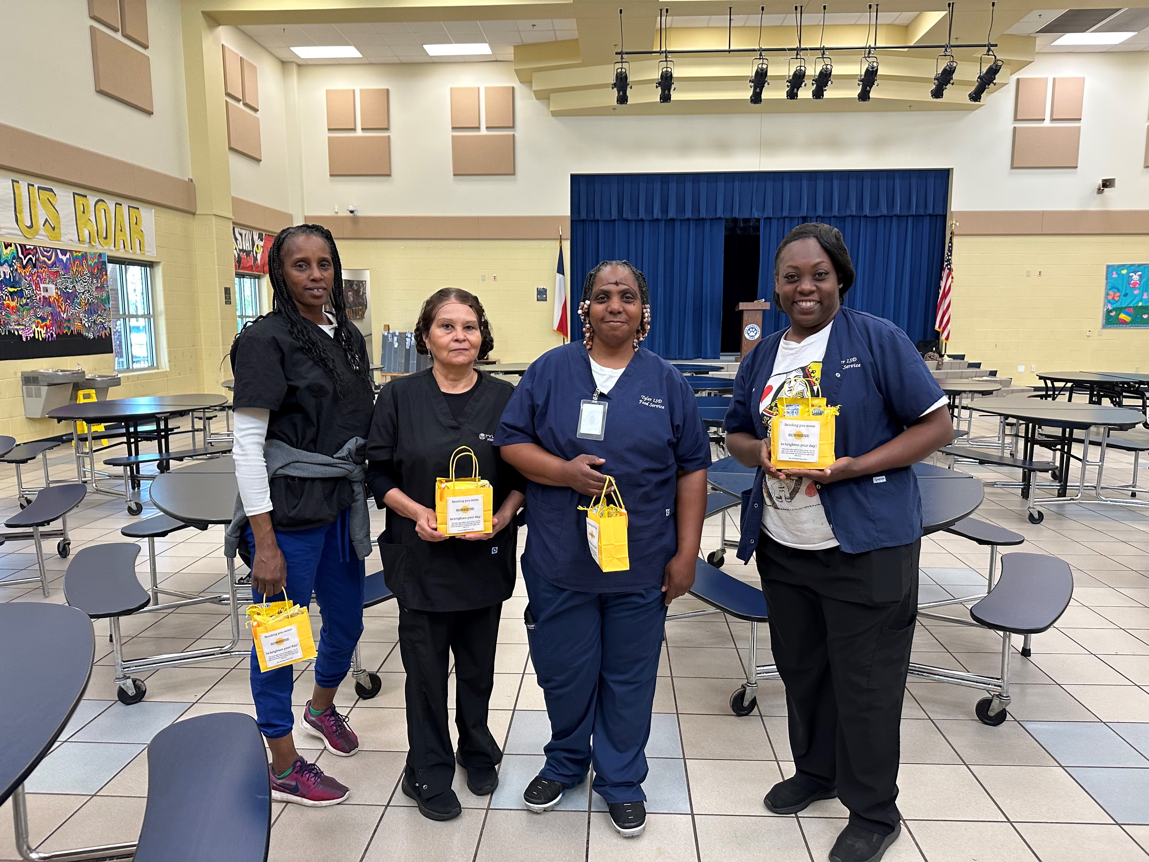 four women standing in a school cafeteria holding yellow gift bags