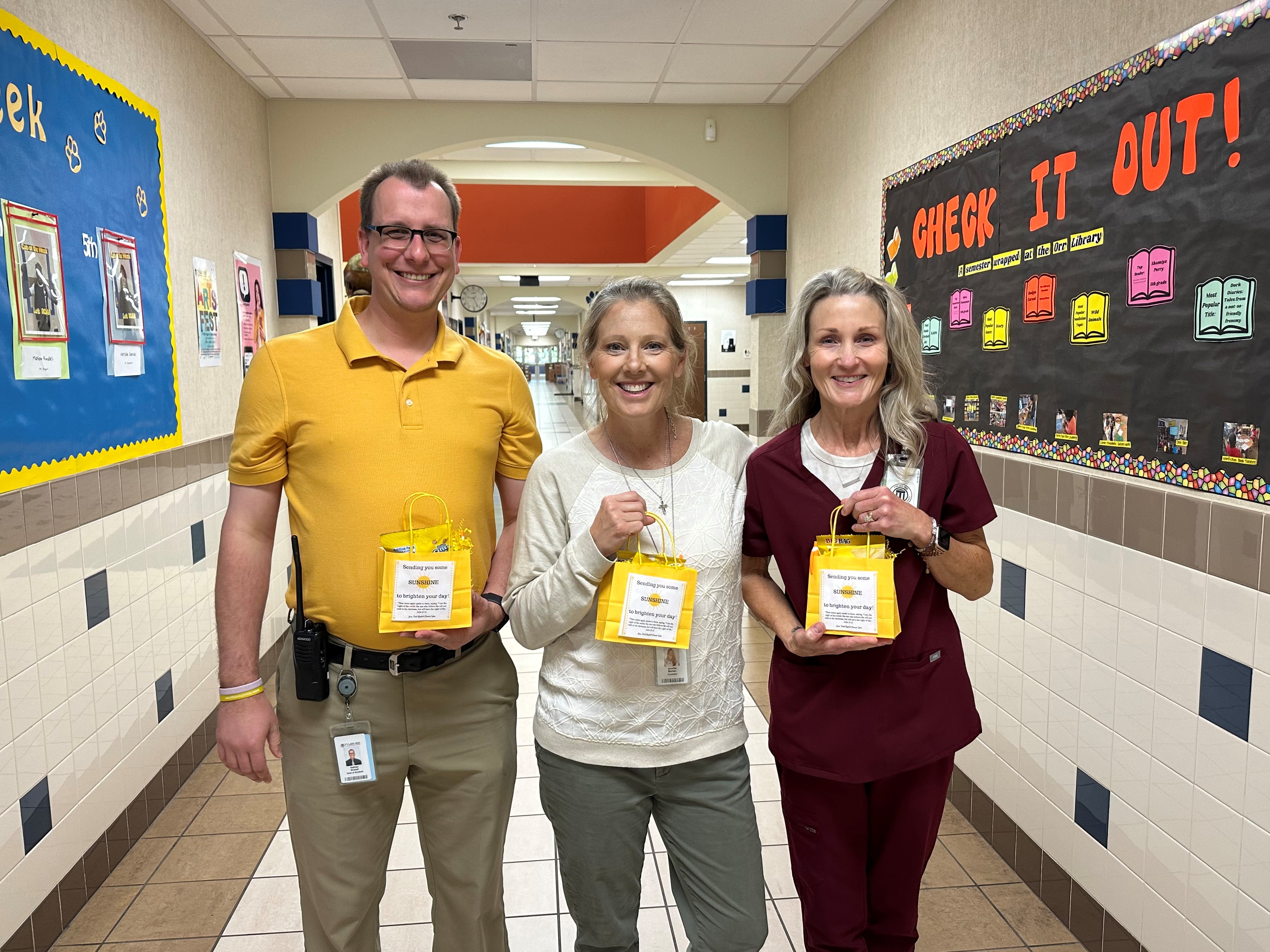 man and two women standing in a school cafeteria holding yellow gift bags