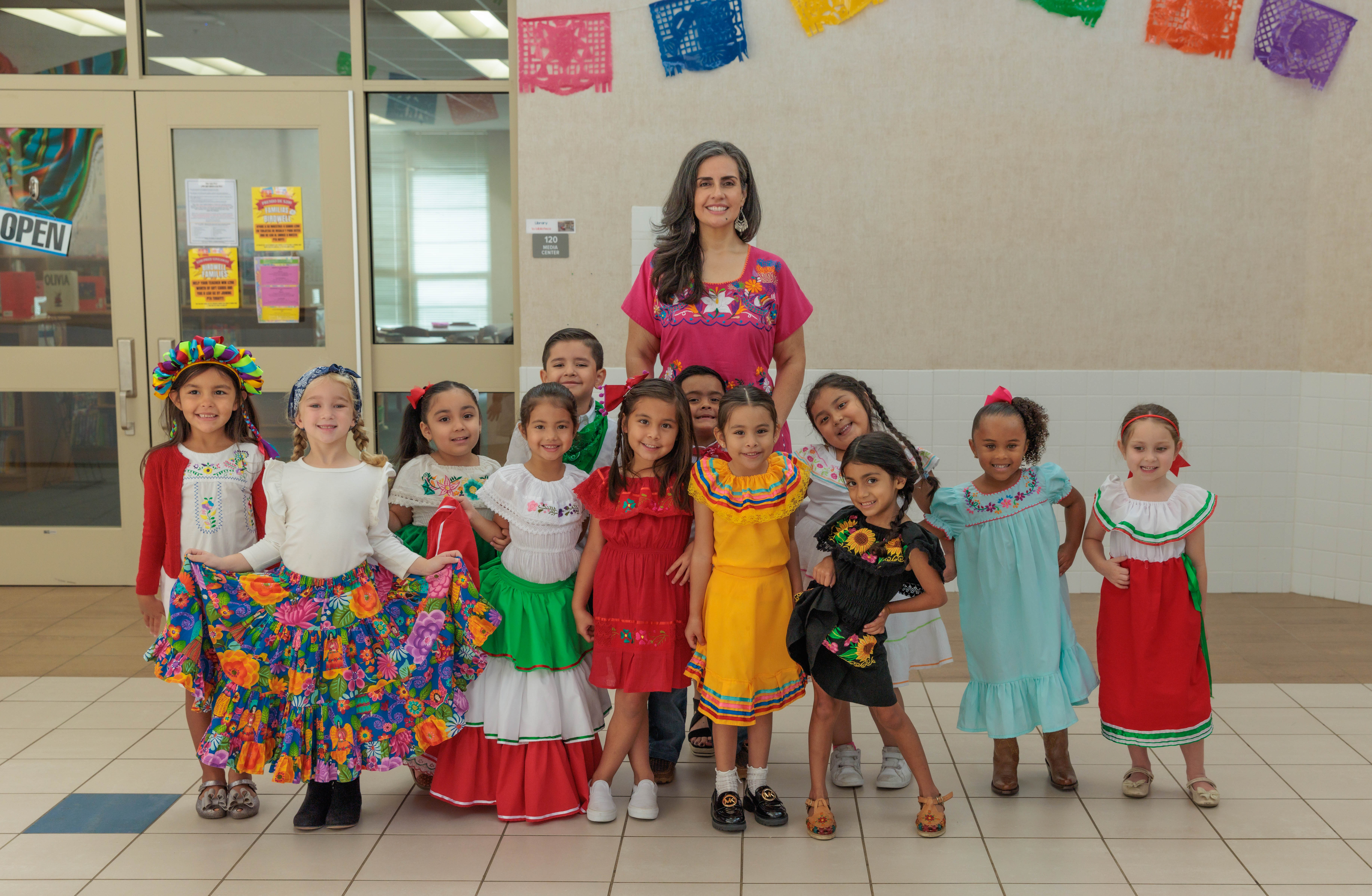 elementary age students and their Hispanic woman principal stand in a group wearing traditional Hispanic clothing