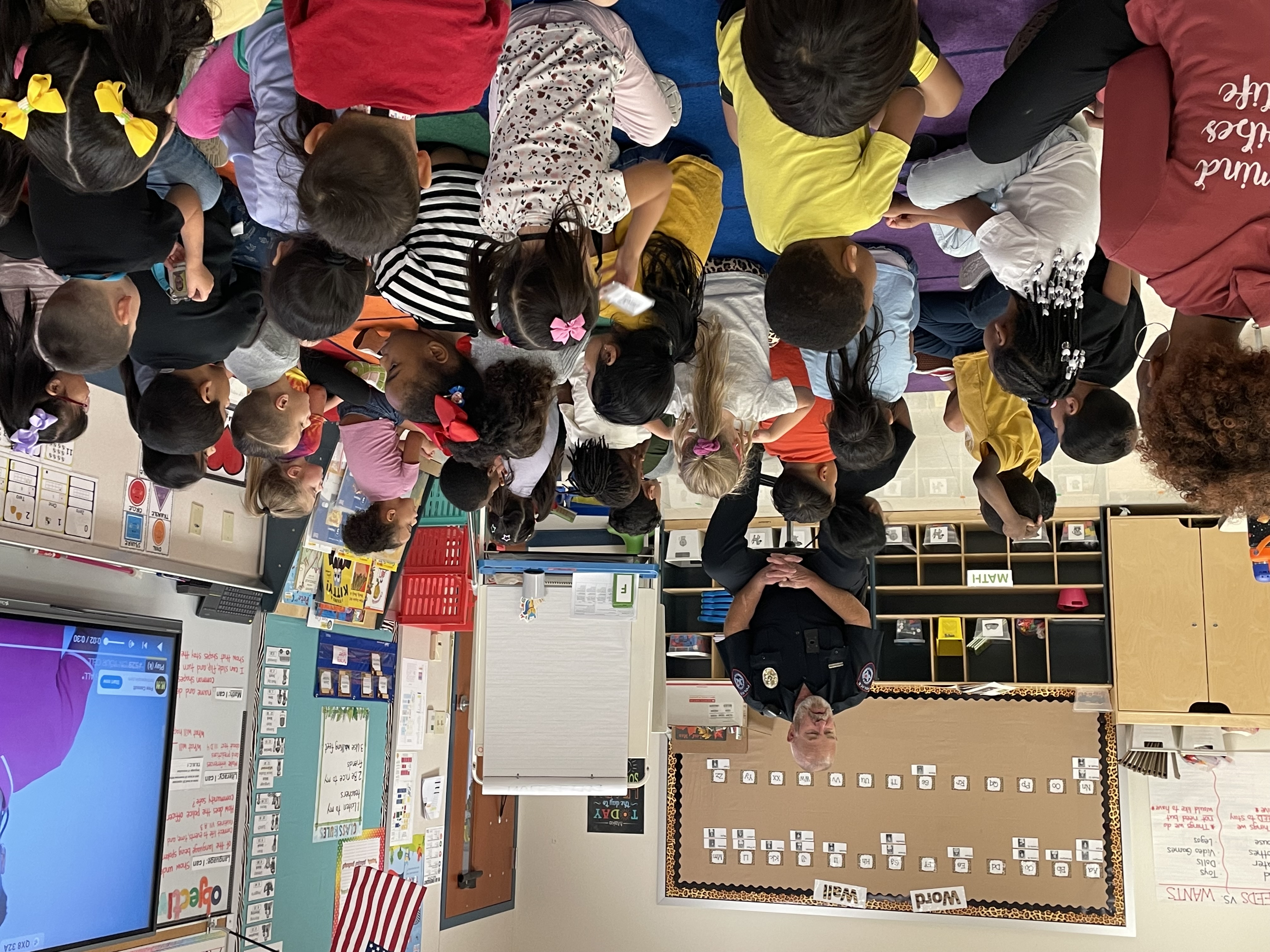 pre-k students sitting on a rug in a classroom with a police officer sitting in a chair talking to them