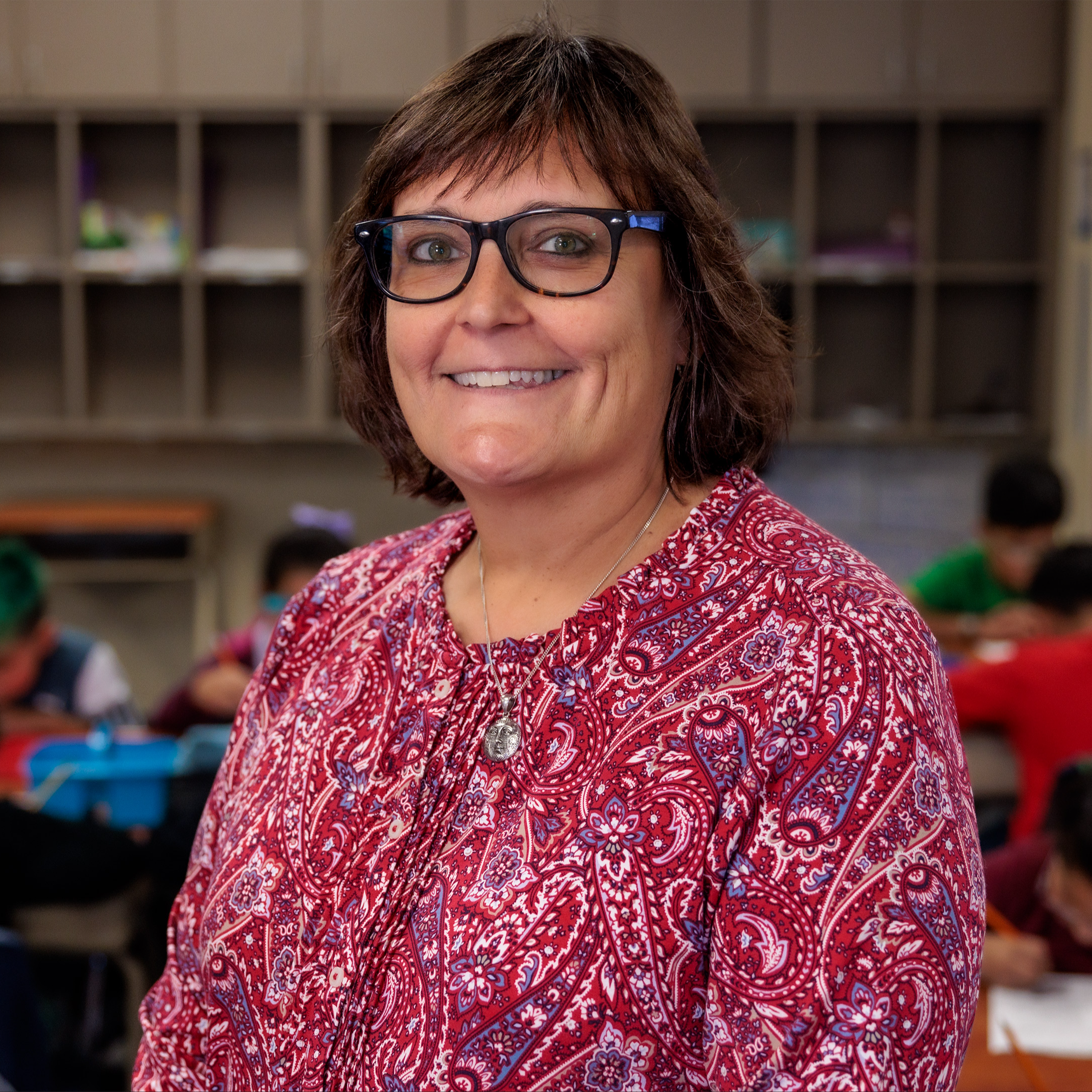 Caucasian woman wearing a red print shirt and glasses standing in her classroom