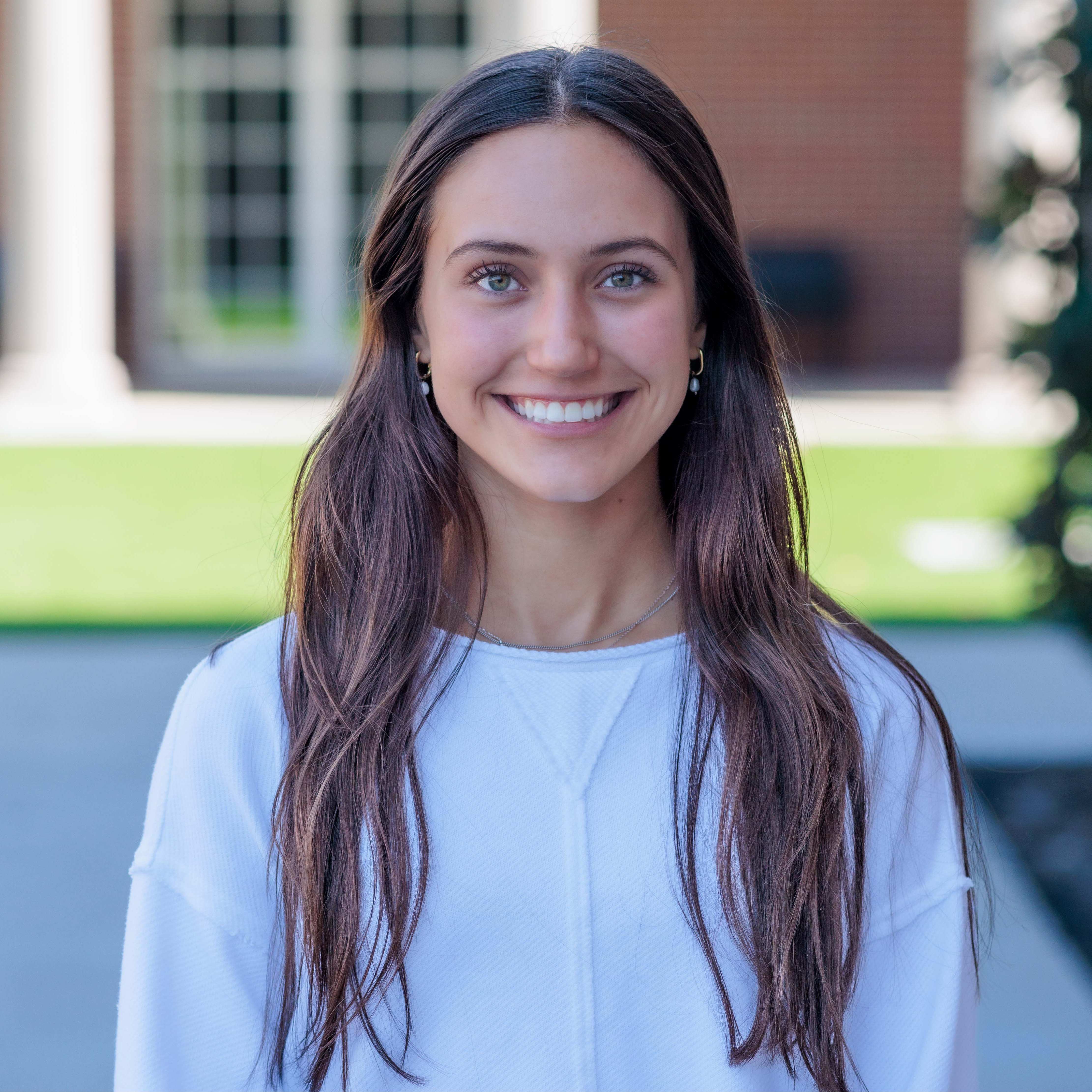 Caucasian high school girl with long brown hair wearing a white top standing outside her school