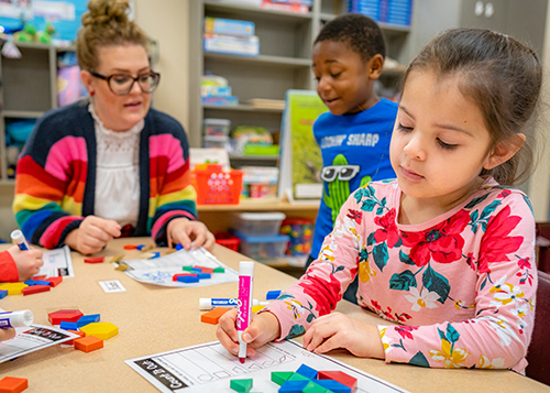 female teacher and two students at a table. female student working on paper with expo markers. bright colors in clothing and markers on table