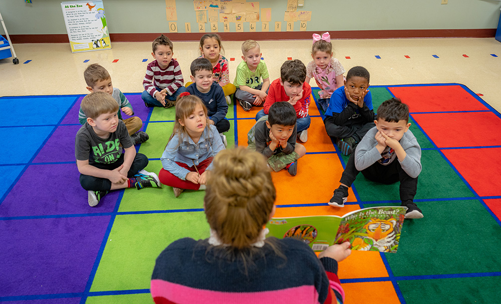 back of a teachers head, with her hair in a bun, reading to a class of kinder students sitting on a rug with blue, purple, green, orange and red squares
