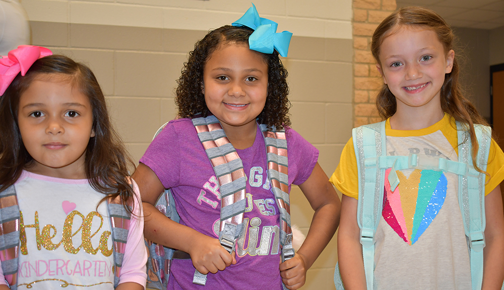 three female kinder students standing next to each other, wearing backpacks. Big smiles on their faces, ready for the first day of school!