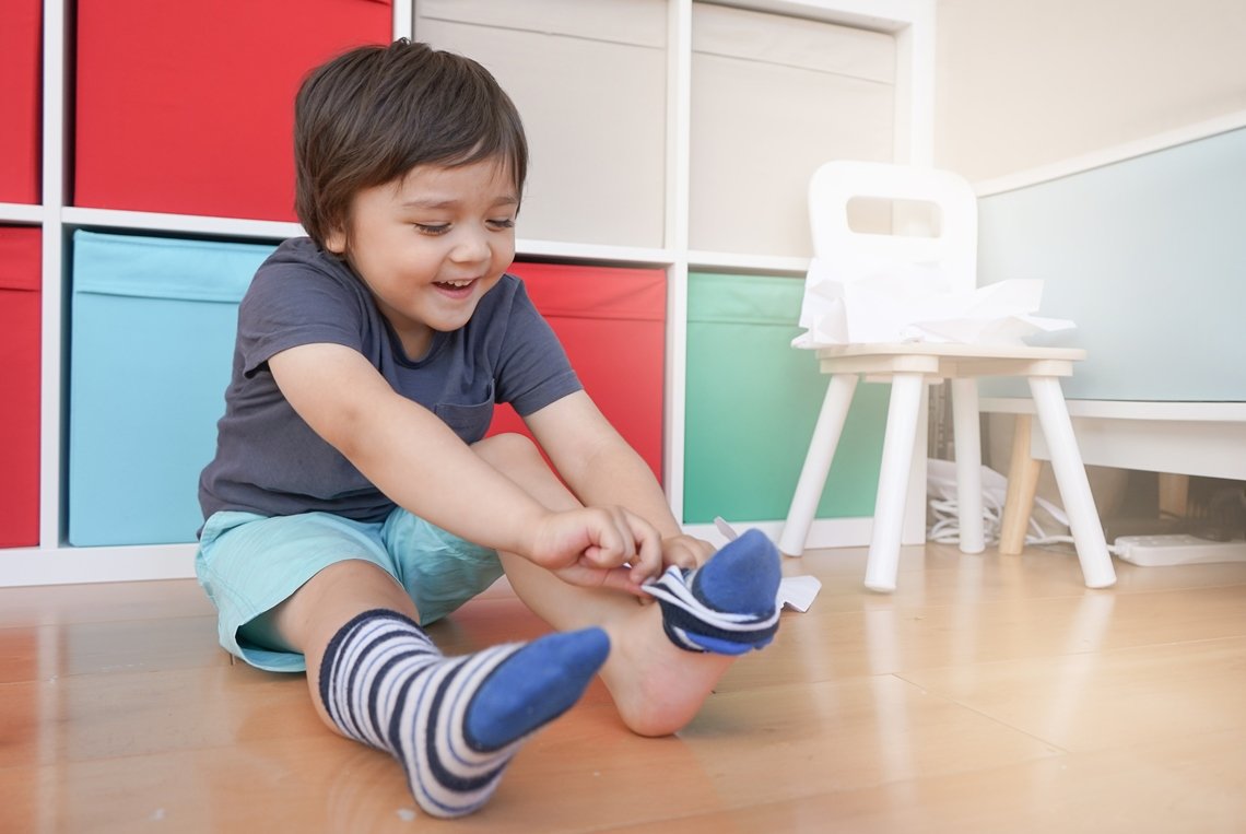 small male child with dark hair, wearing a gray t-shirt and turquoise shorts, sitting on the hardwood floor putting his striped blue socks on