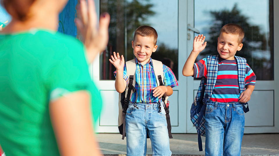 two young boys standing in front of a school door wearing backpacks and waving goodbye to someone