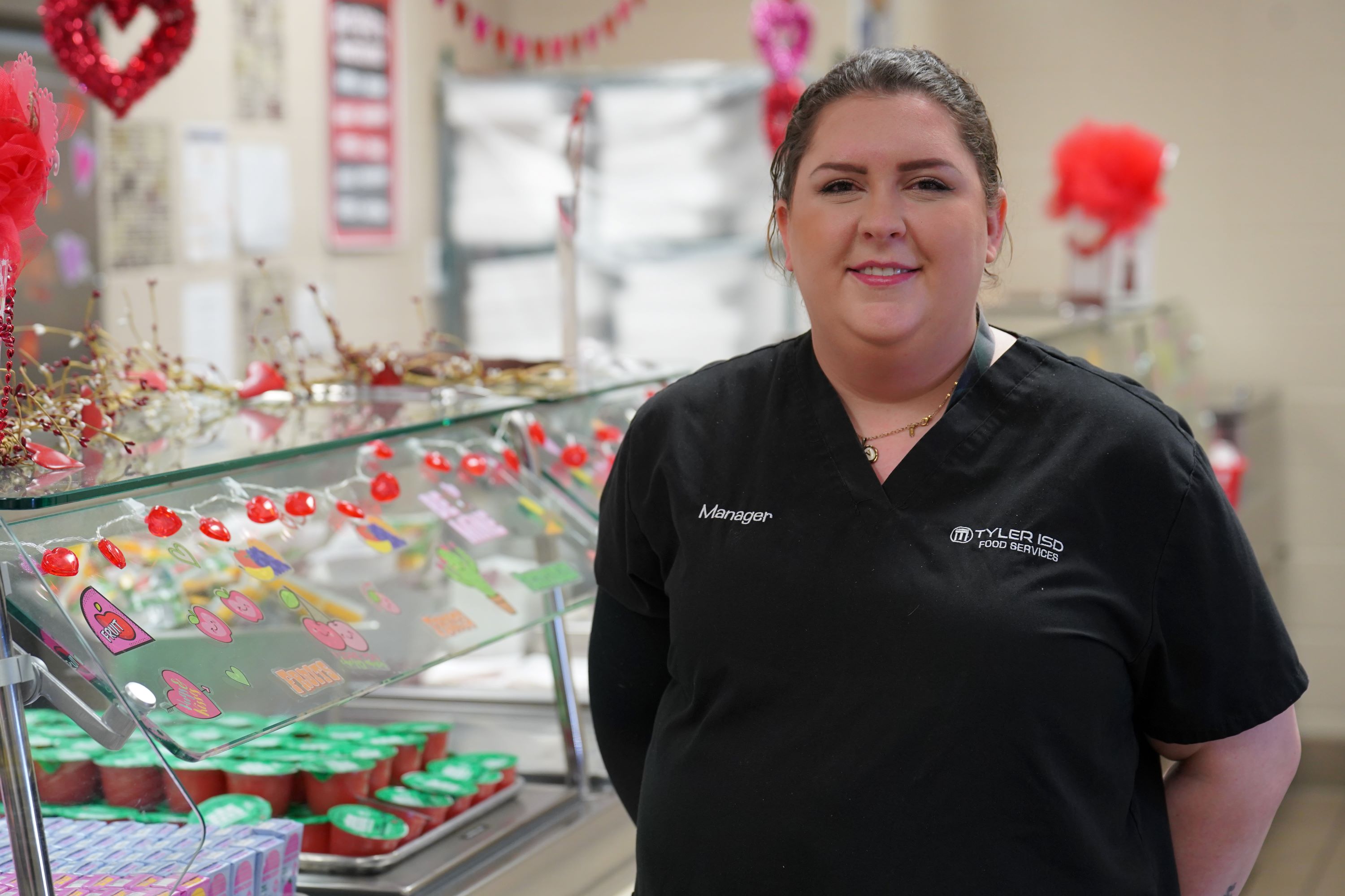 Caucasian woman wearing black scrubs standing in front of a cafeteria food line