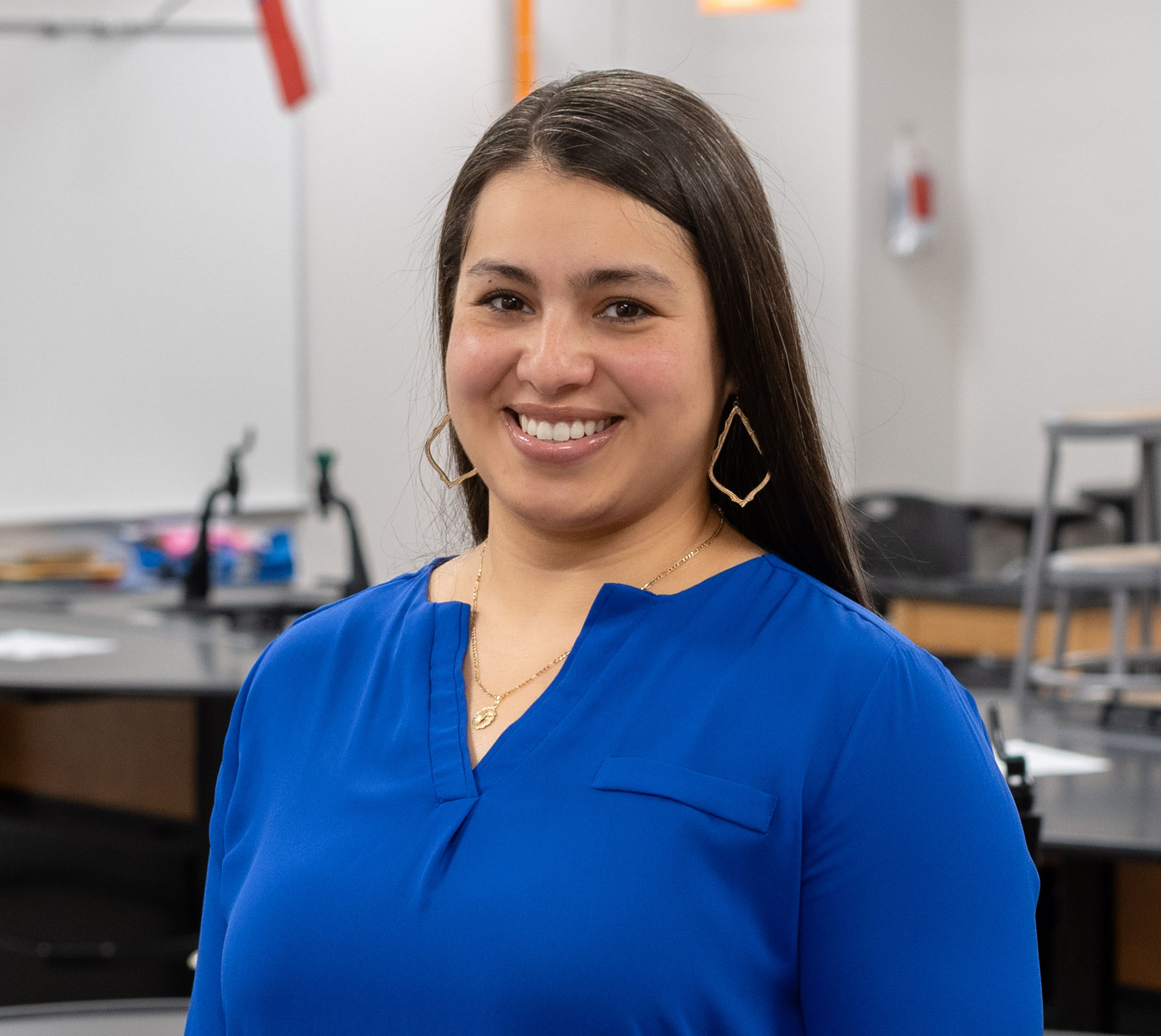 woman with long dark hair wearing a blue blouse standing in her classroom