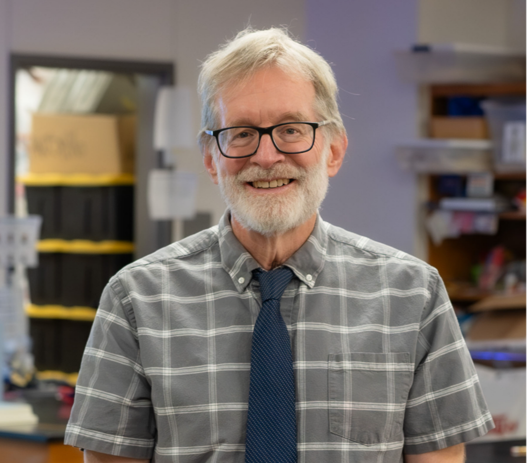 Caucasian man with gray hair and glasses wearing a gray plaid shirt and blue tie