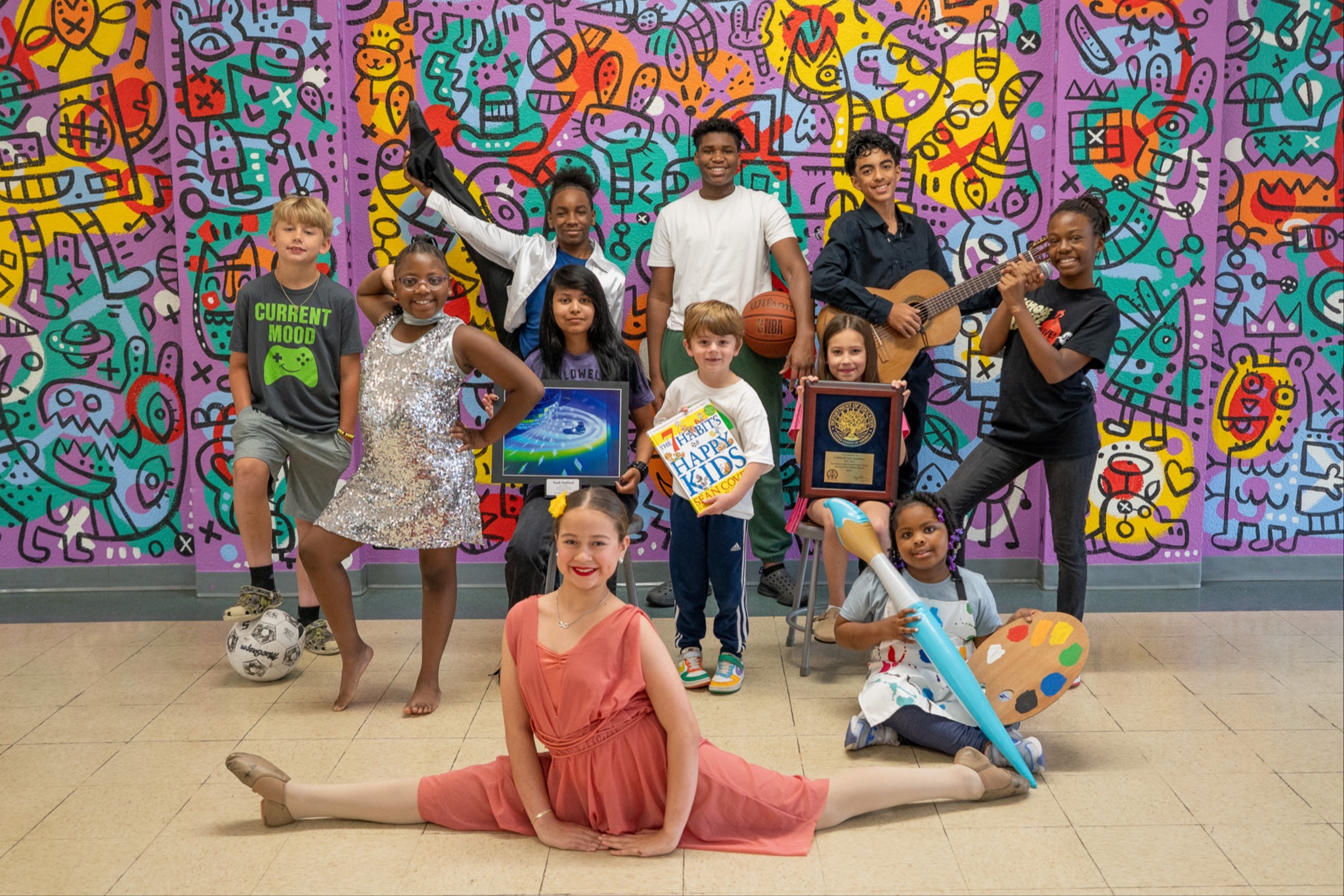 A group of children poses in front of a brightly colored, graffiti-style mural. They hold various objects.